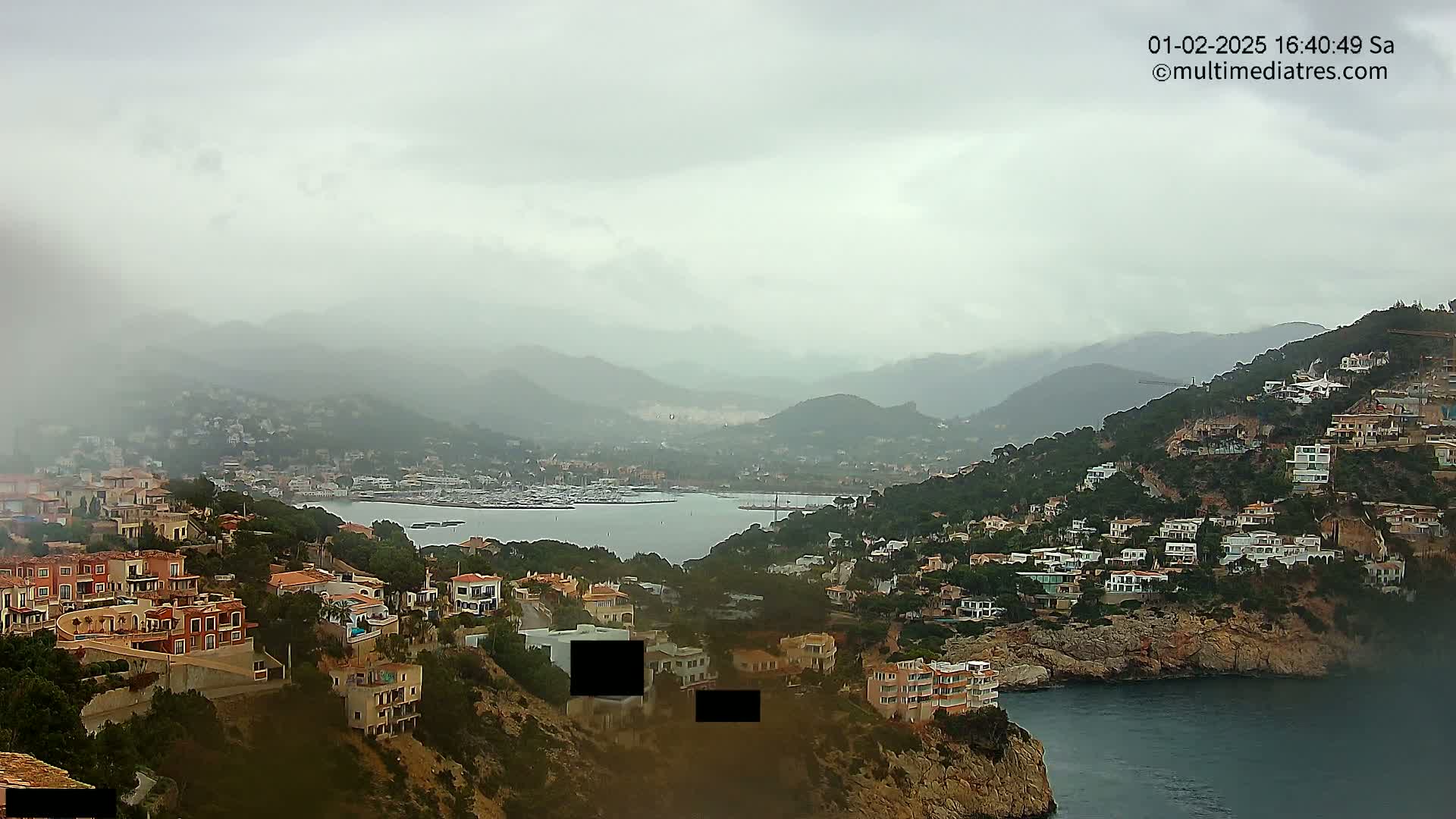 A coastal town nestled in a hilly landscape under a cloudy, overcast sky overlooks a calm bay filled with boats.