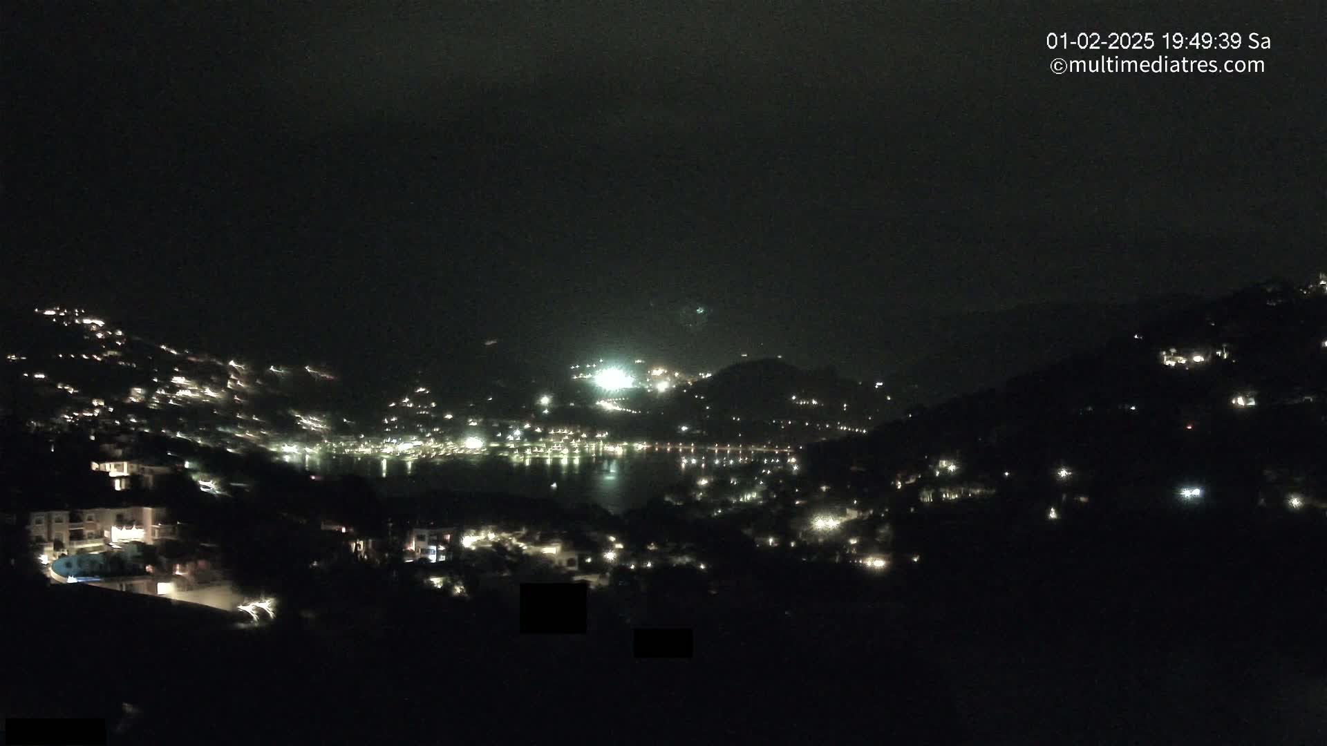 A nighttime view of a coastal town illuminated by scattered lights, with a dark body of water in the distance under a dark, possibly cloudy sky.