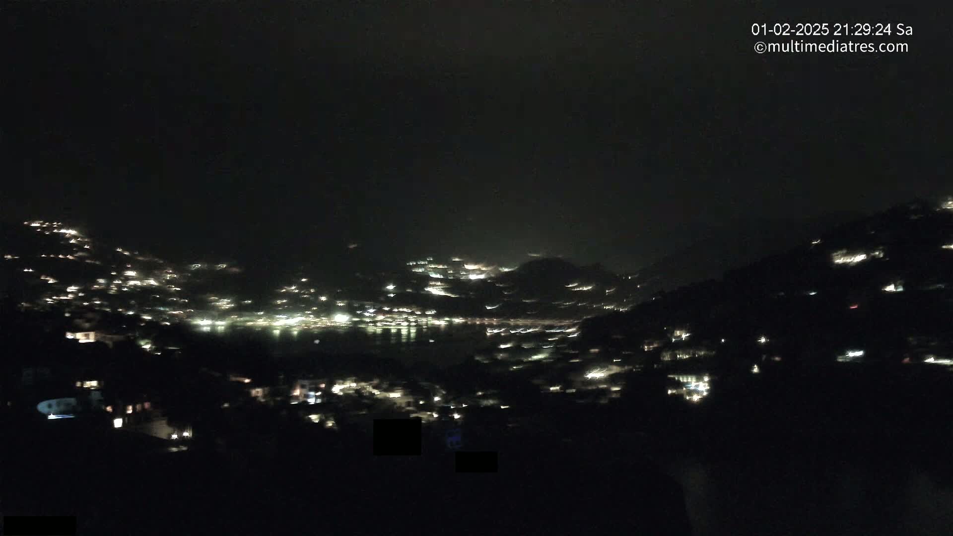 A nighttime view shows a dark, hilly landscape with scattered lights from buildings and a body of water reflecting light in the distance under a dark, cloudy sky.