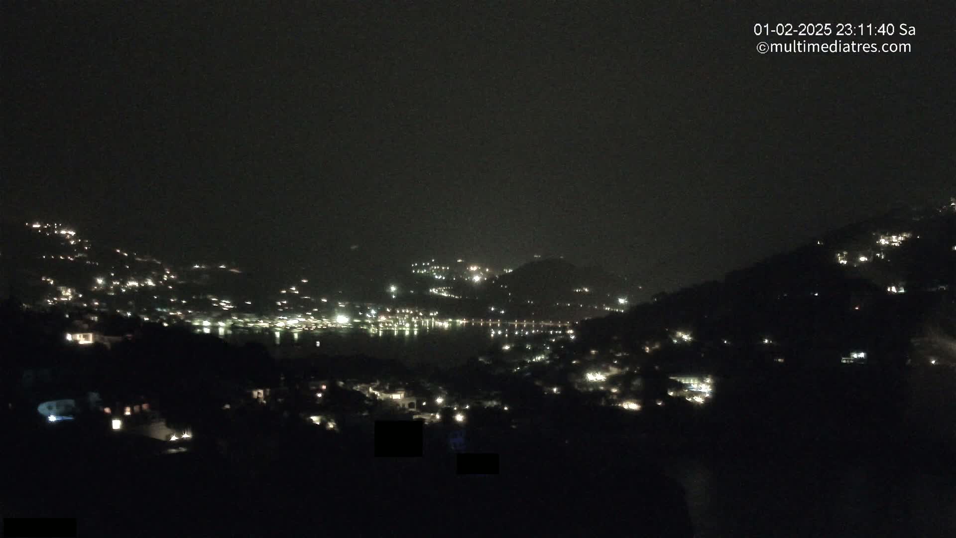 A nighttime view of a coastal town nestled in hills, with scattered lights illuminating the landscape under a dark, cloudy sky.
