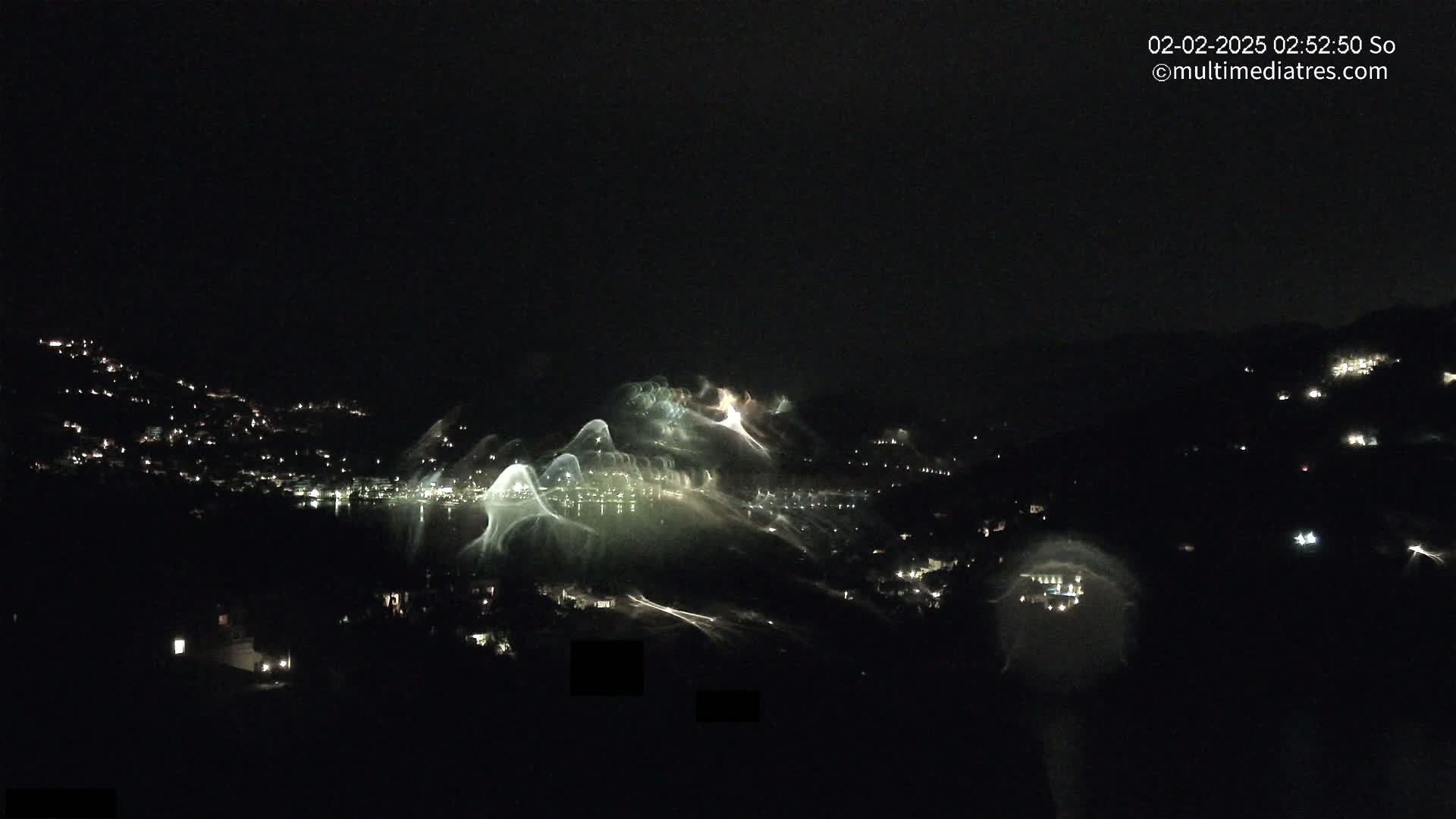 A nighttime view of a snow-covered hillside with illuminated ski runs and a town with scattered lights below.