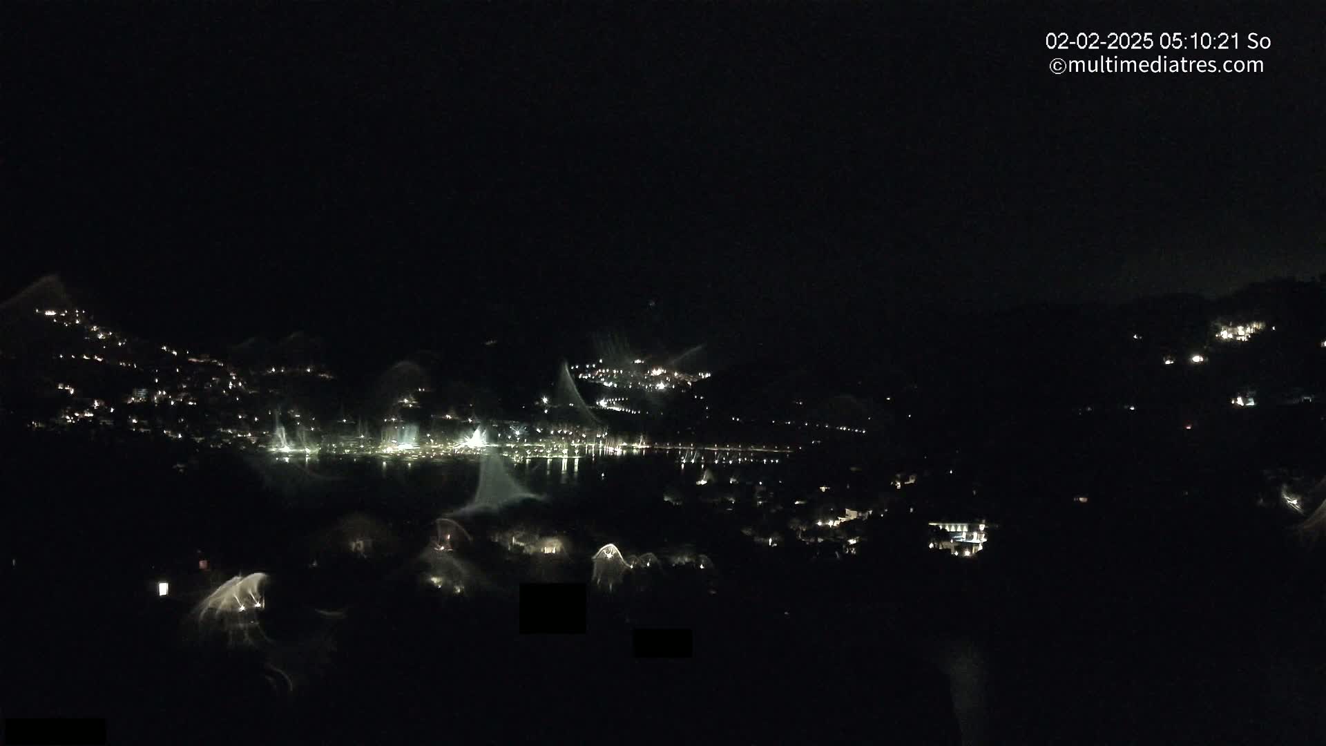 A nighttime view shows a body of water with lights reflecting on it, surrounded by hills dotted with lights, under a clear, dark sky.