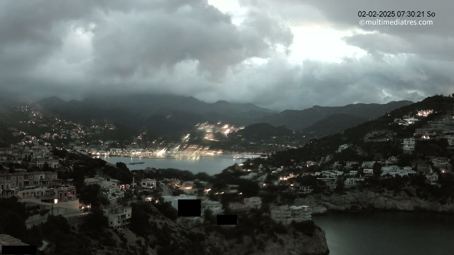 A nighttime view of a coastal town nestled between dark mountains under a cloudy sky.