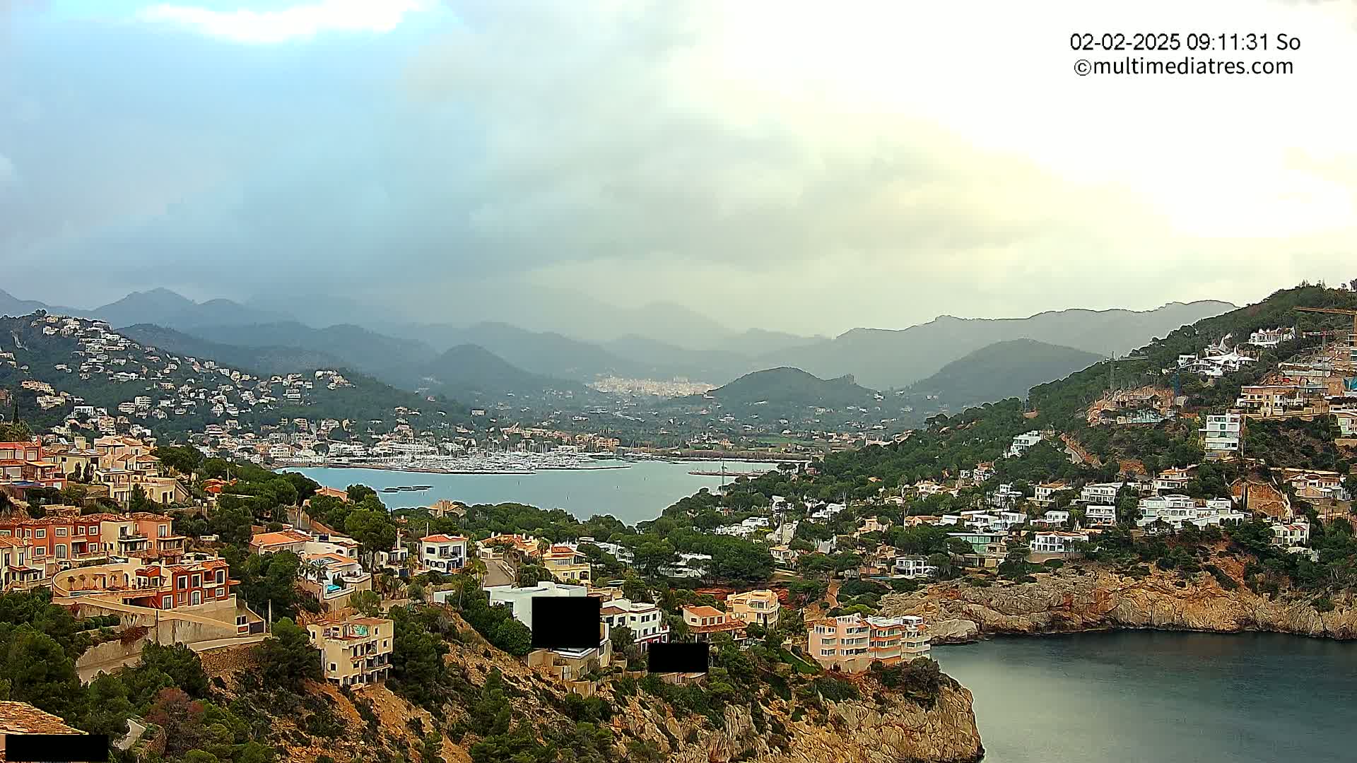 A coastal town nestled in hills overlooks a calm bay filled with boats under a cloudy sky.