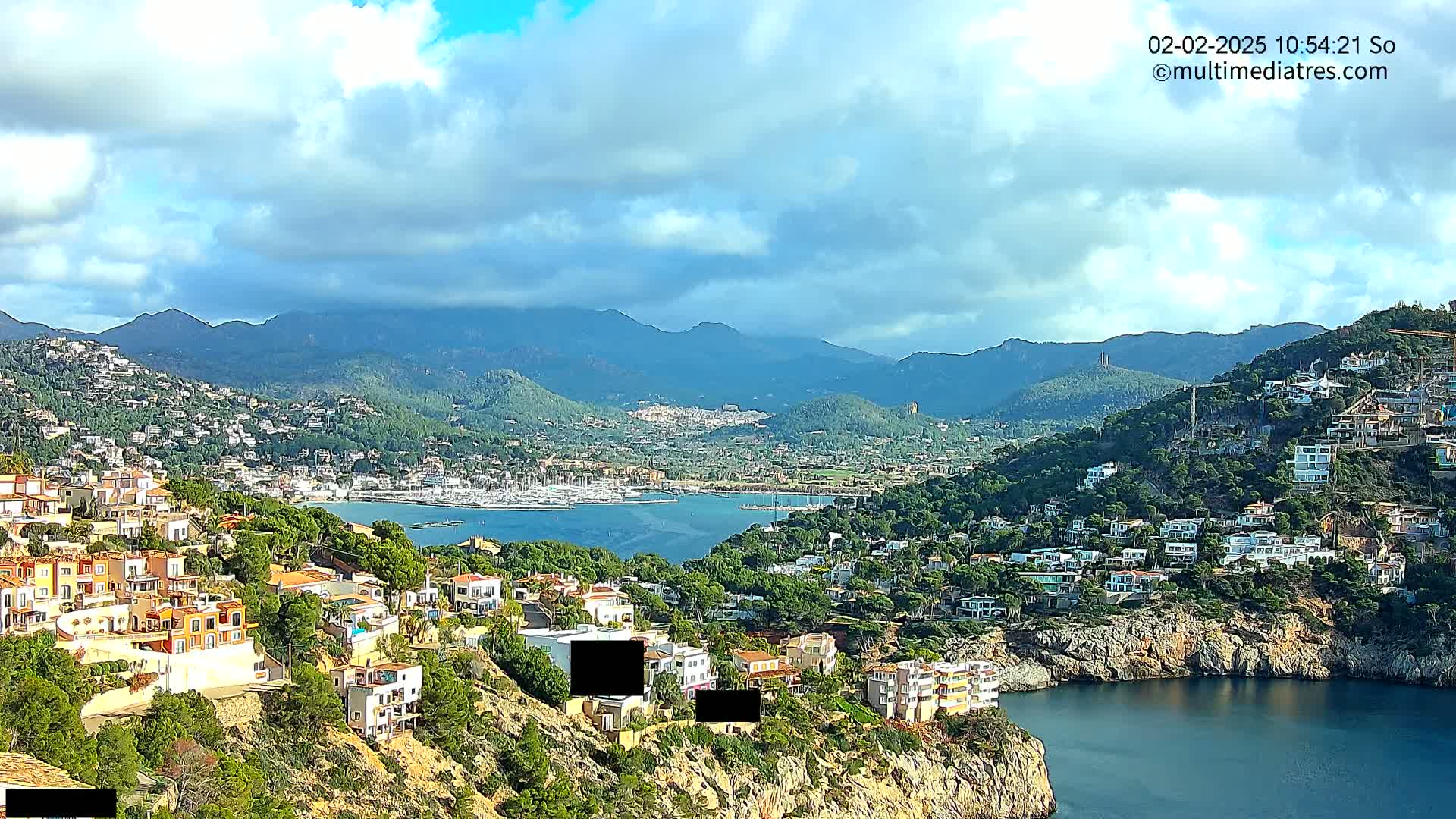 A coastal town nestled on a hillside overlooks a calm bay filled with boats, with mountains in the background under a partly cloudy sky.