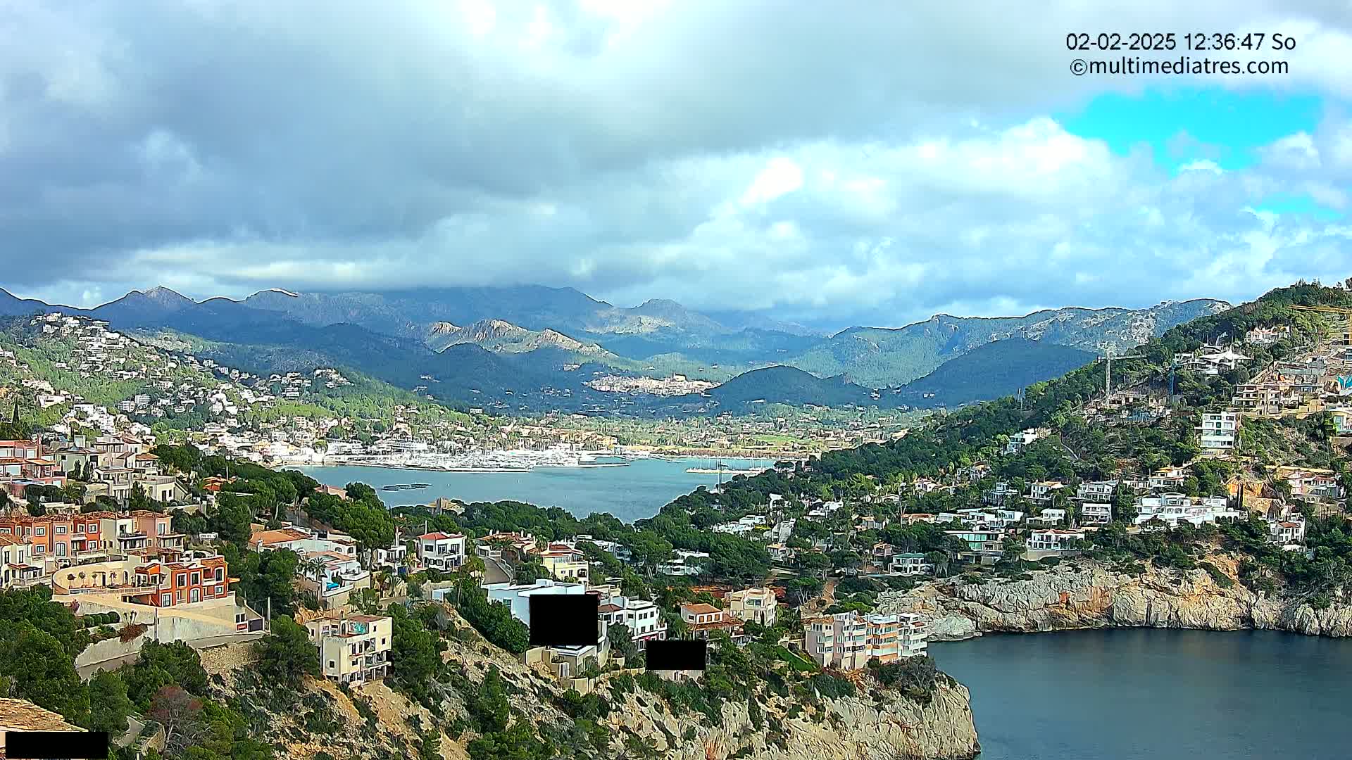 A coastal town nestled in a mountainous region under a partly cloudy sky overlooks a calm bay filled with boats.