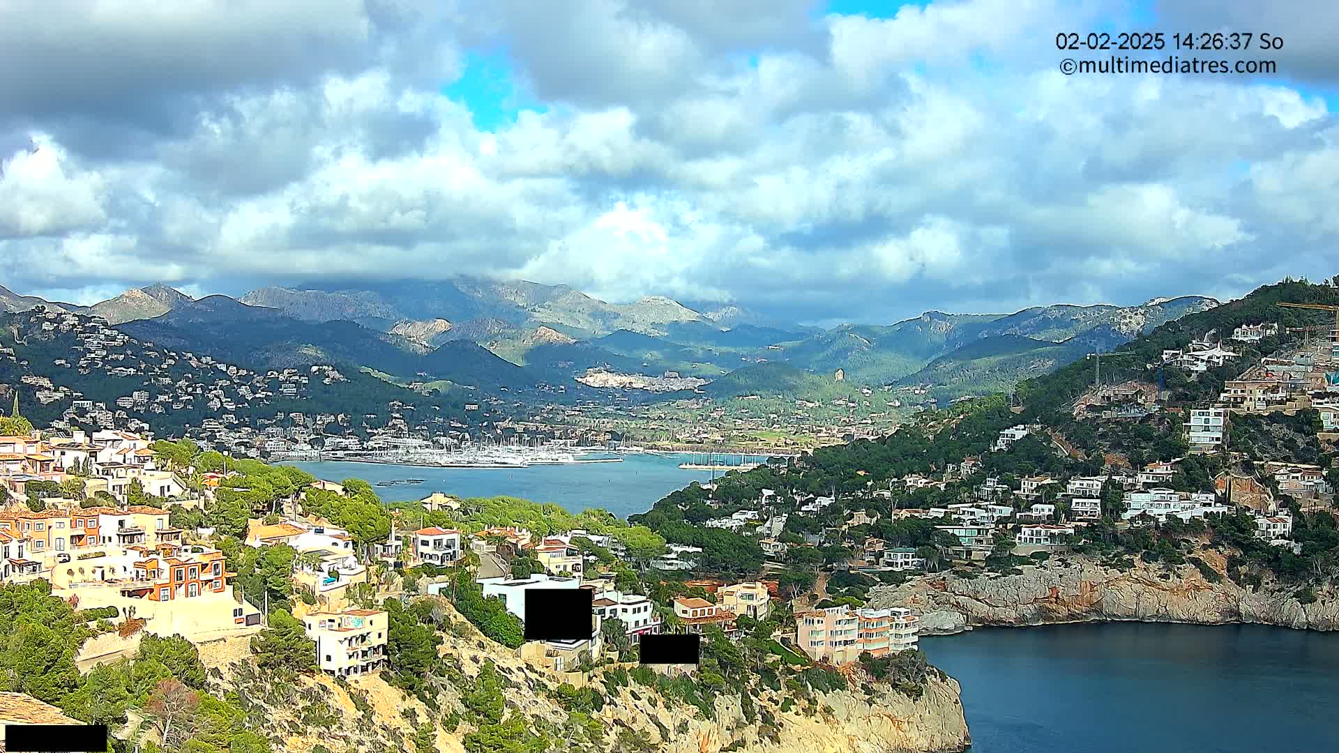 A coastal town nestled in a mountainous region under a partly cloudy sky, with a calm bay filled with boats visible in the distance.
