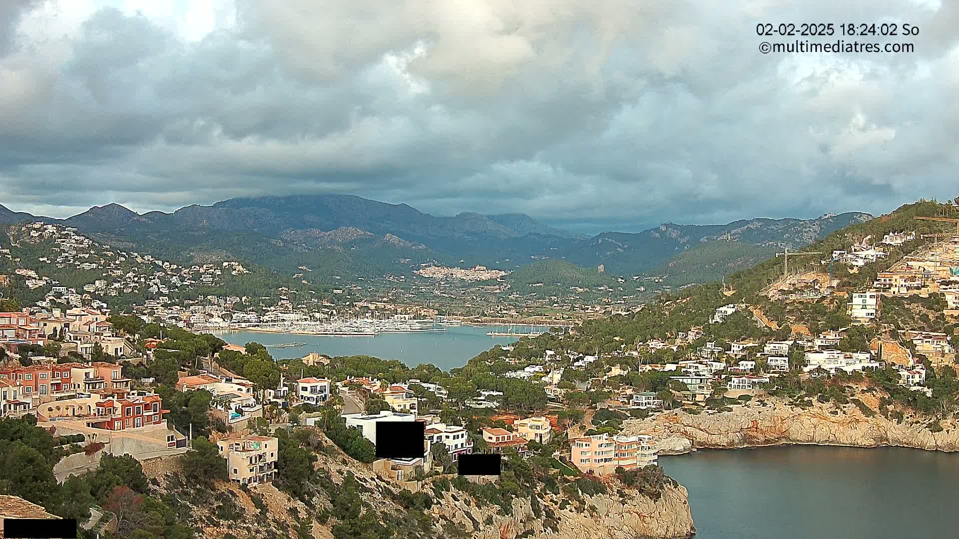 A coastal town nestled in a mountainous region under a cloudy sky overlooks a calm bay filled with boats.