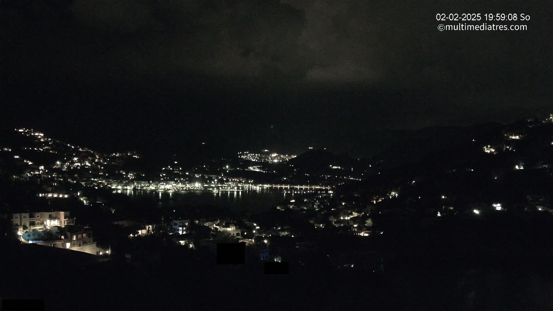 A nighttime view of a coastal town illuminated by scattered lights, under a dark, cloudy sky.