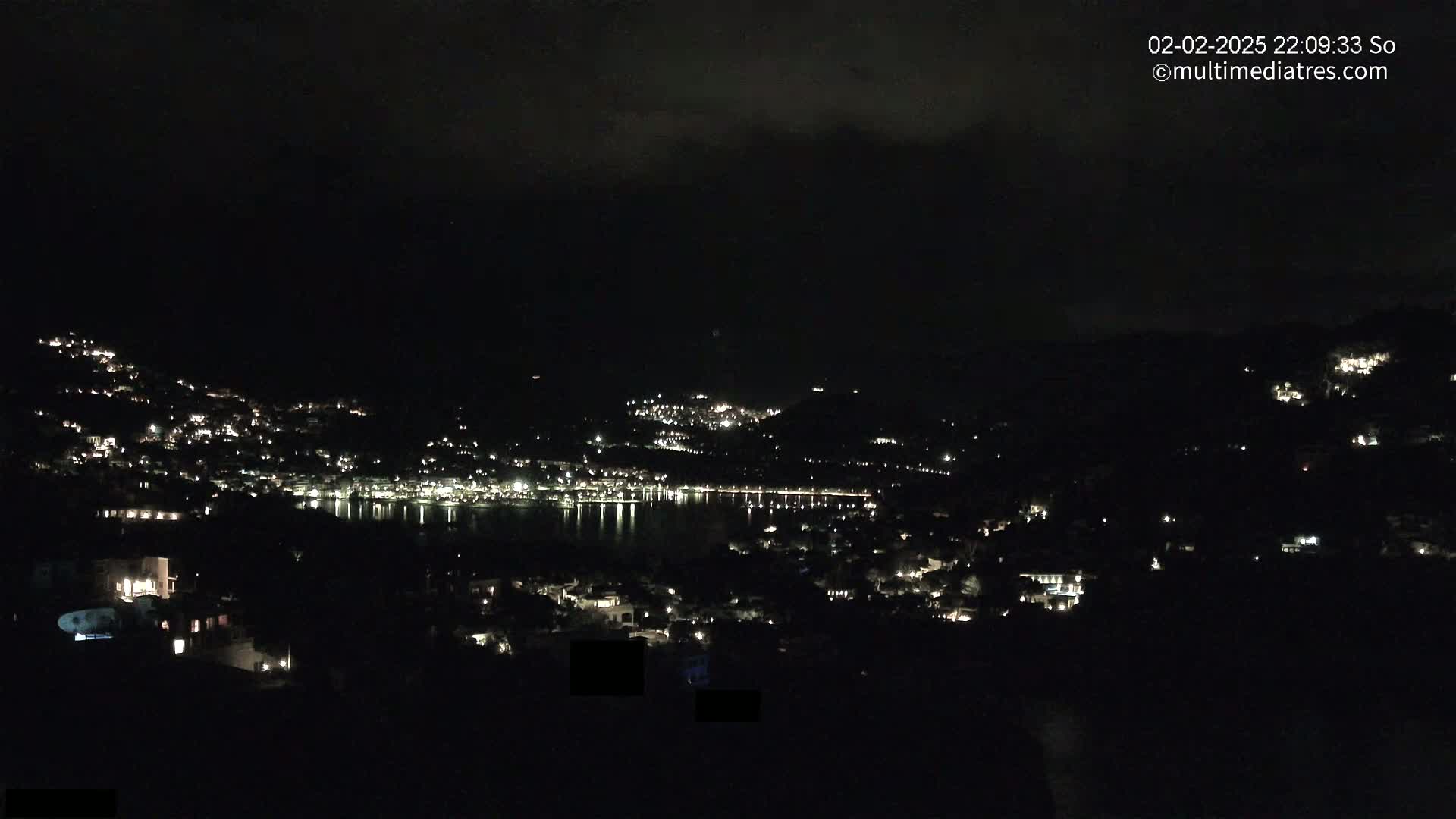 A nighttime view of a coastal town with scattered lights on hillsides surrounding a dark body of water under a cloudy sky.