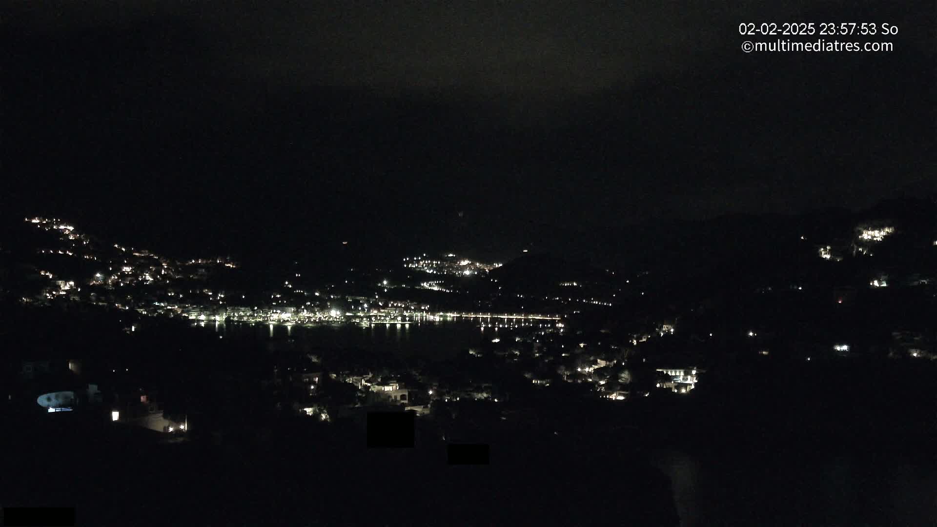 A nighttime view shows a town nestled on hillsides surrounding a dark body of water under a cloudy sky.