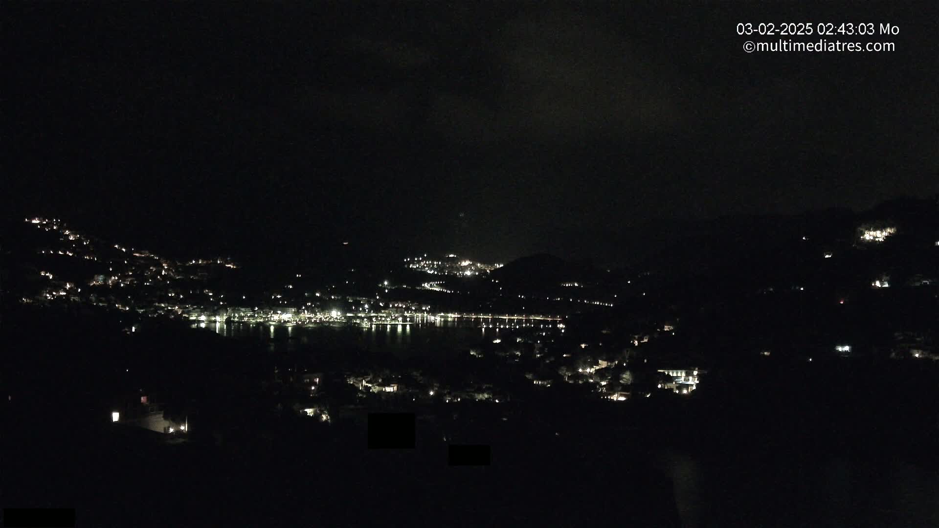 A nighttime view shows a town nestled on hillsides surrounding a dark body of water, with scattered lights illuminating the landscape under a mostly clear sky.