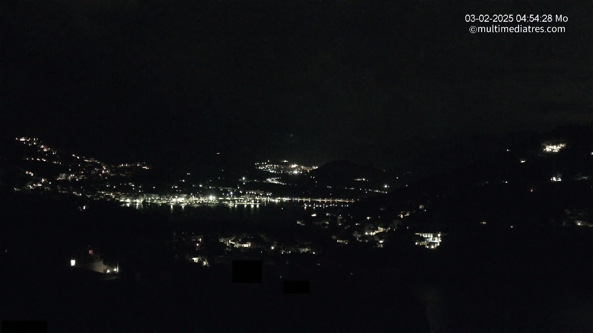 A nighttime view of a town nestled on hillsides around a dark body of water, under a clear, dark sky.