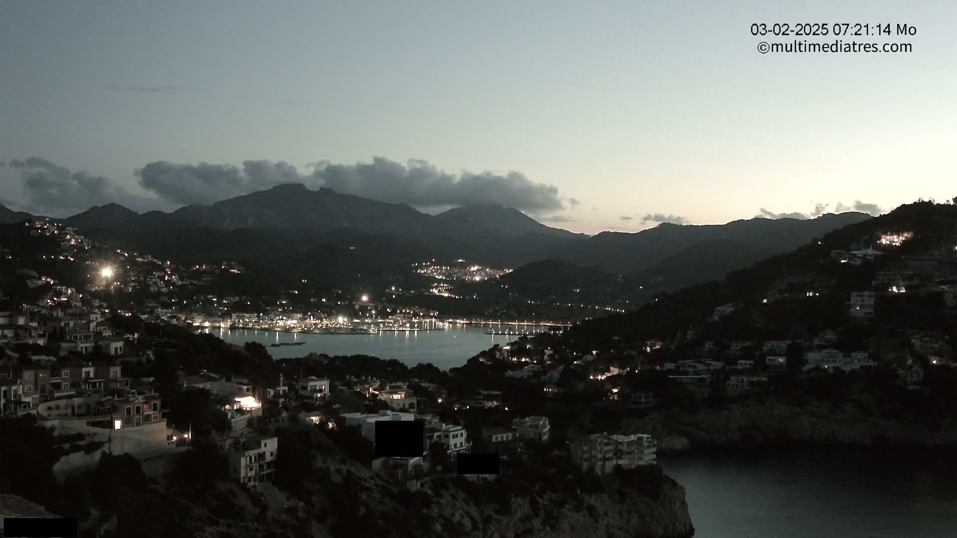 A coastal town nestled between dark mountains is illuminated at twilight under a mostly cloudy sky.