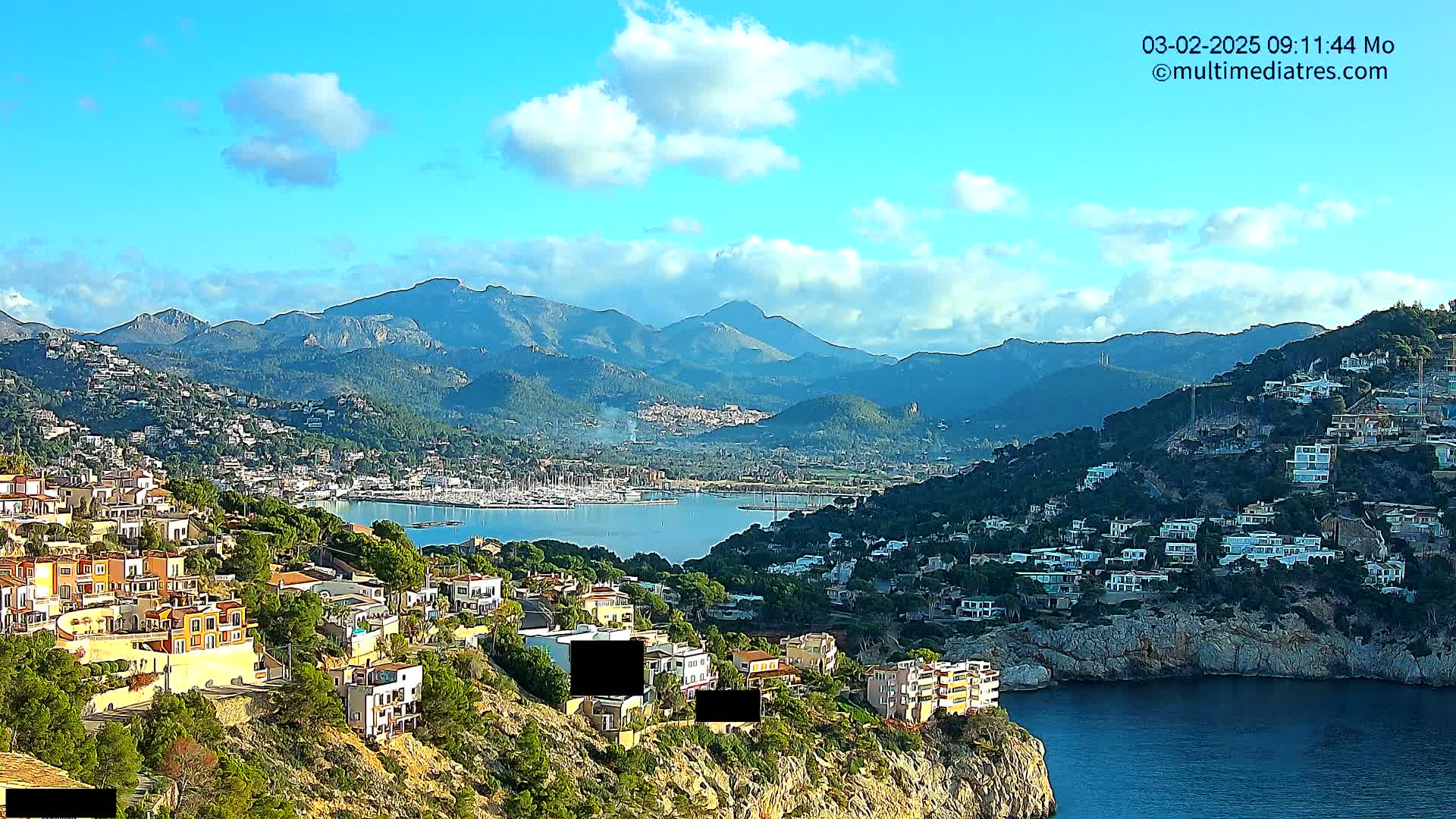 A coastal town nestled in hills overlooking a calm bay filled with boats, with mountains in the background under a mostly sunny sky.