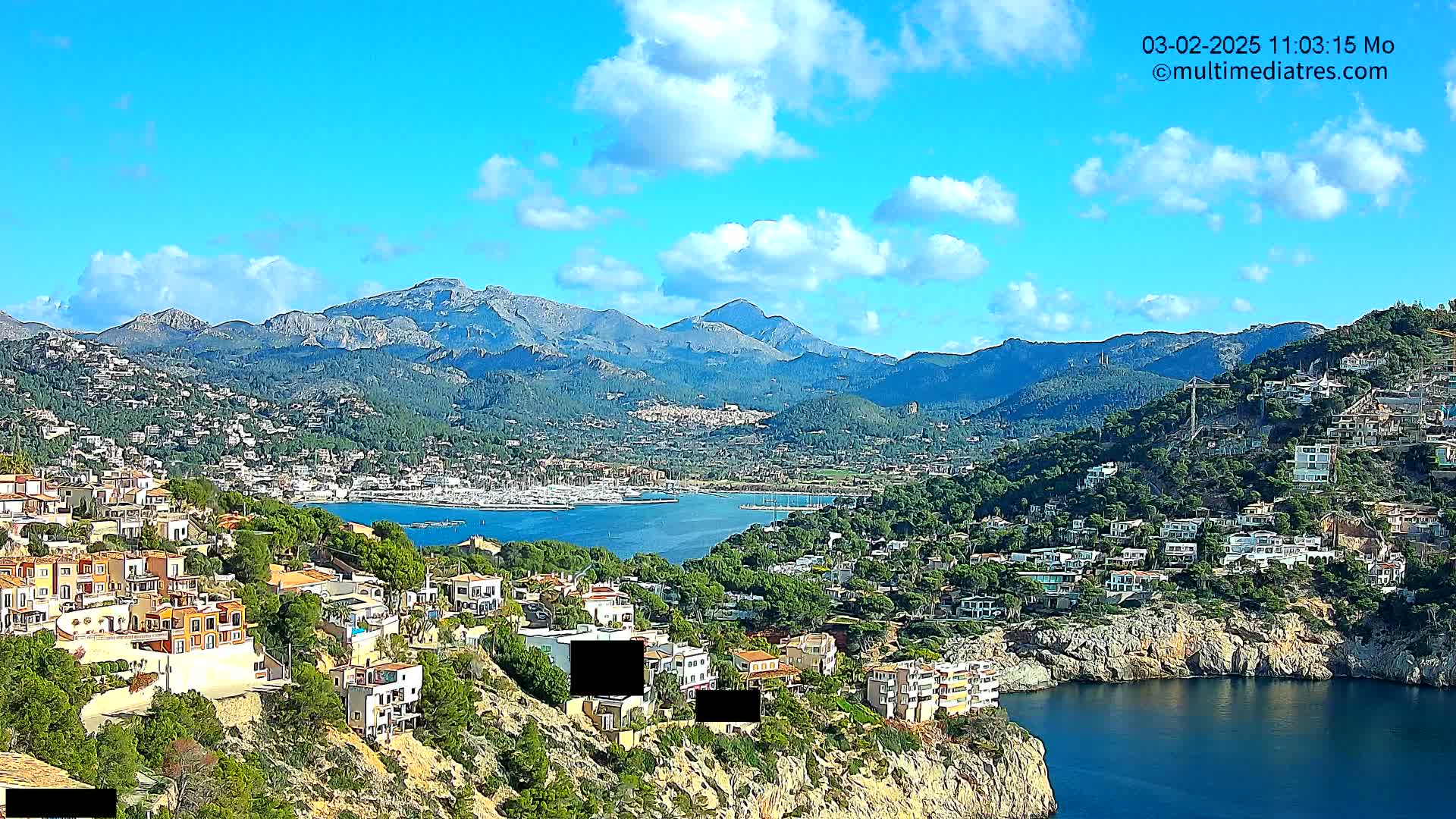 A coastal town nestled between mountains and a calm bay under a partly cloudy blue sky.