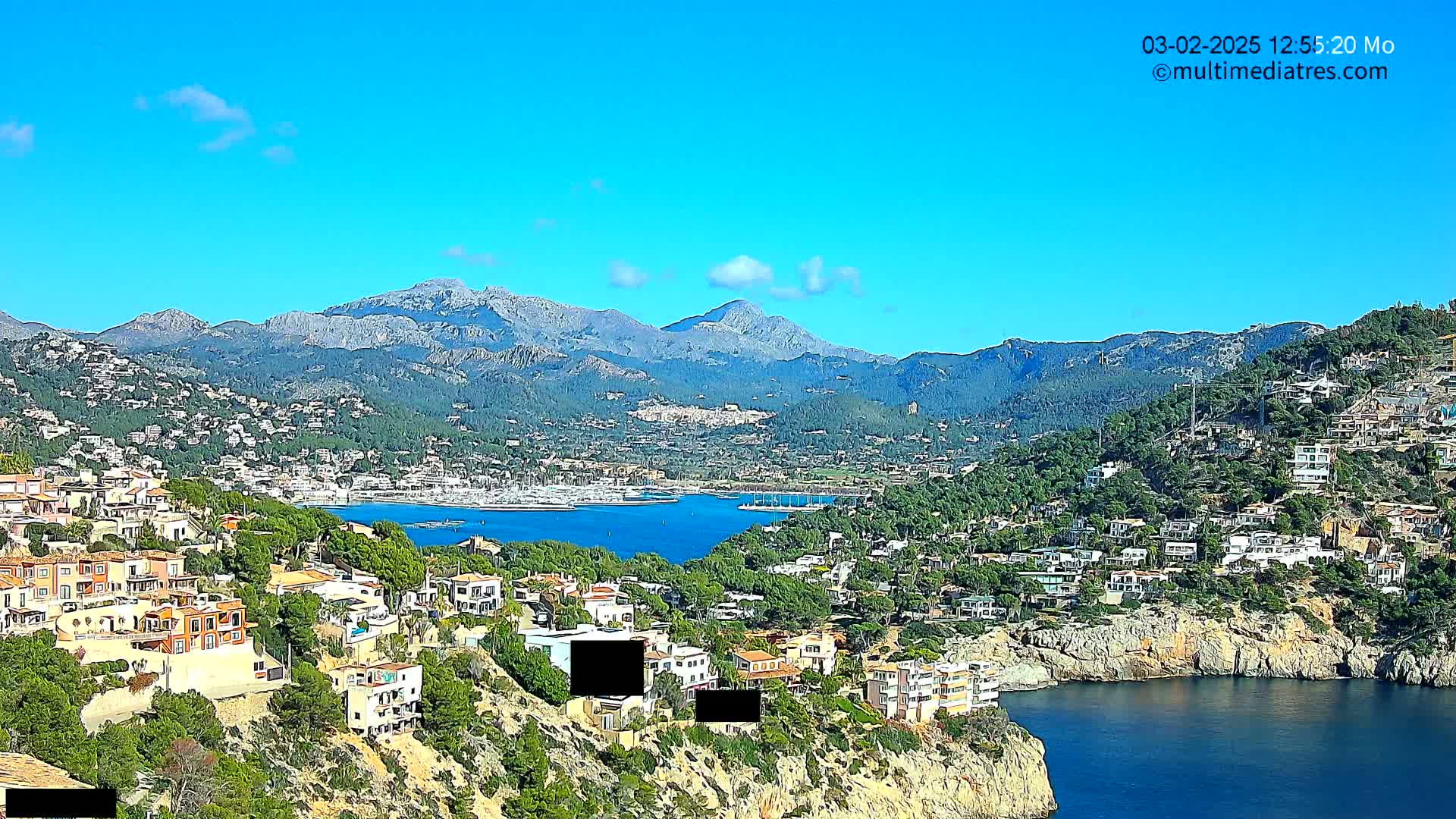 A picturesque coastal town nestled between a calm blue bay and a mountain range under a clear blue sky.