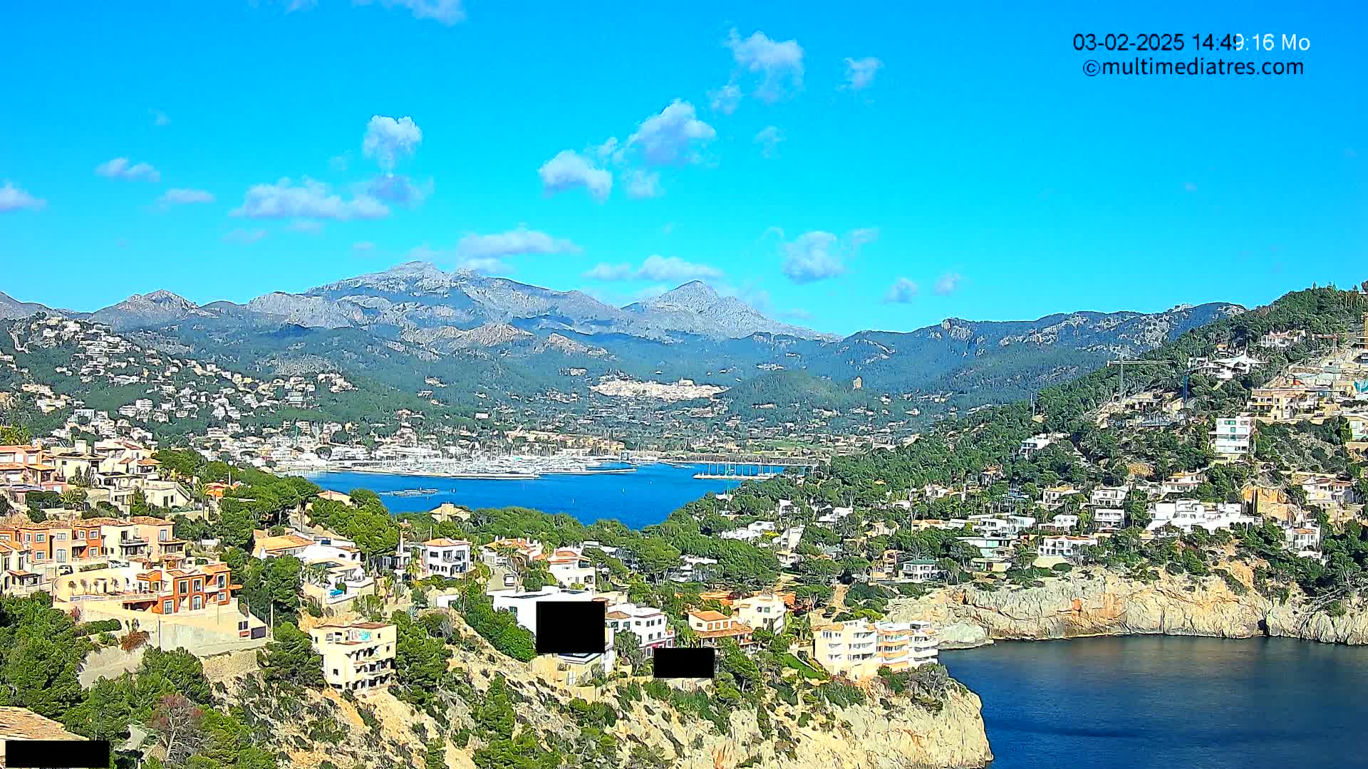 A coastal town nestled on a hillside overlooking a calm bay, with mountains in the background under a clear blue sky with a few clouds.