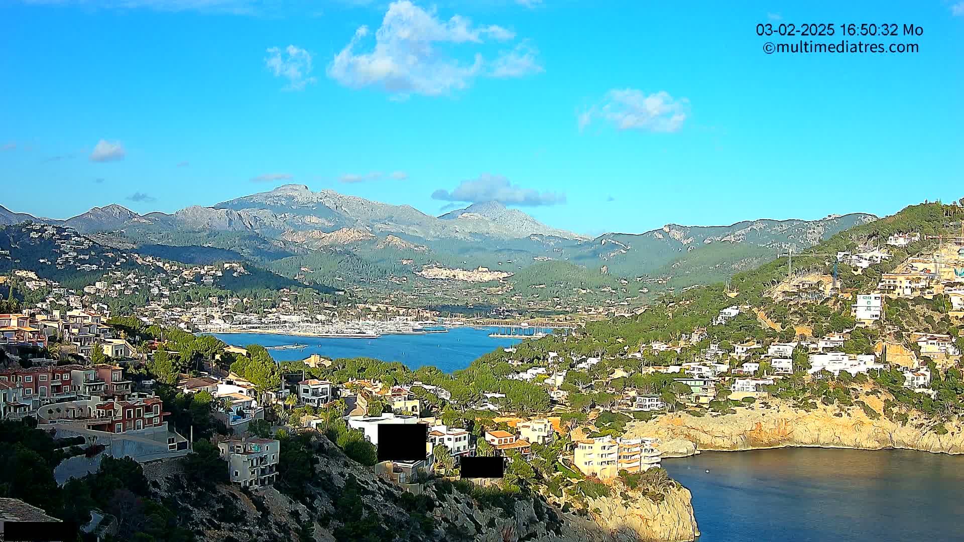 A sunny coastal town nestled in a mountainous region overlooks a calm bay filled with boats, under a mostly clear sky with a few clouds.