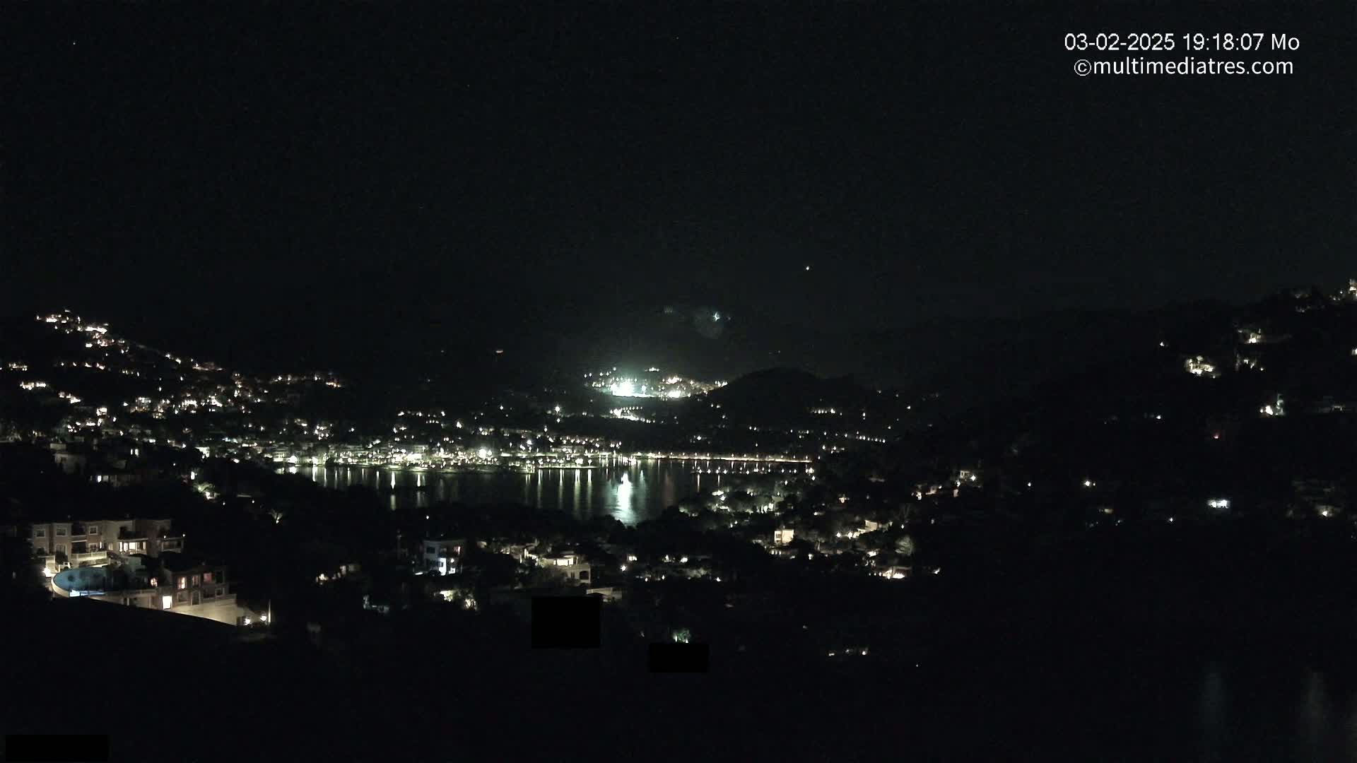 A nighttime view of a coastal town illuminated by scattered lights, nestled between hills and overlooking a calm body of water.