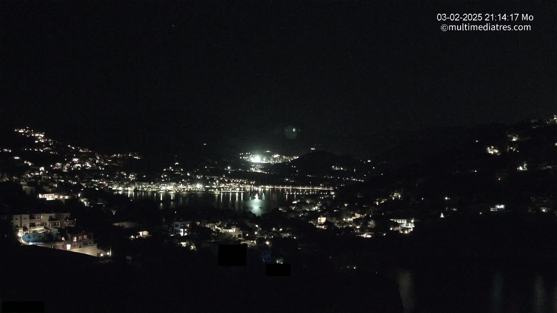 A nighttime view shows a coastal town with many lights illuminating houses on hillsides surrounding a dark body of water.