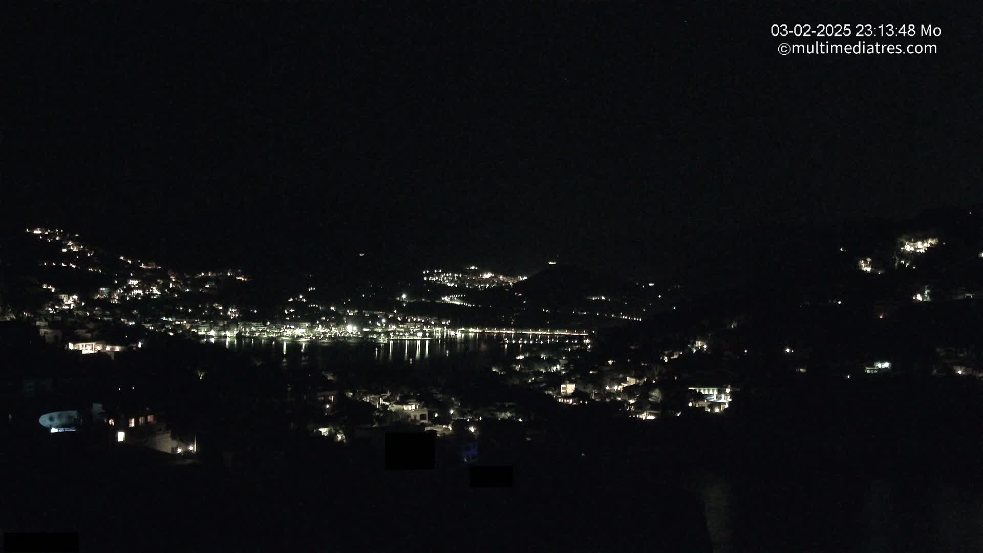A nighttime view of a town nestled on a hillside, illuminated by scattered lights, overlooking a calm body of water reflecting the lights.