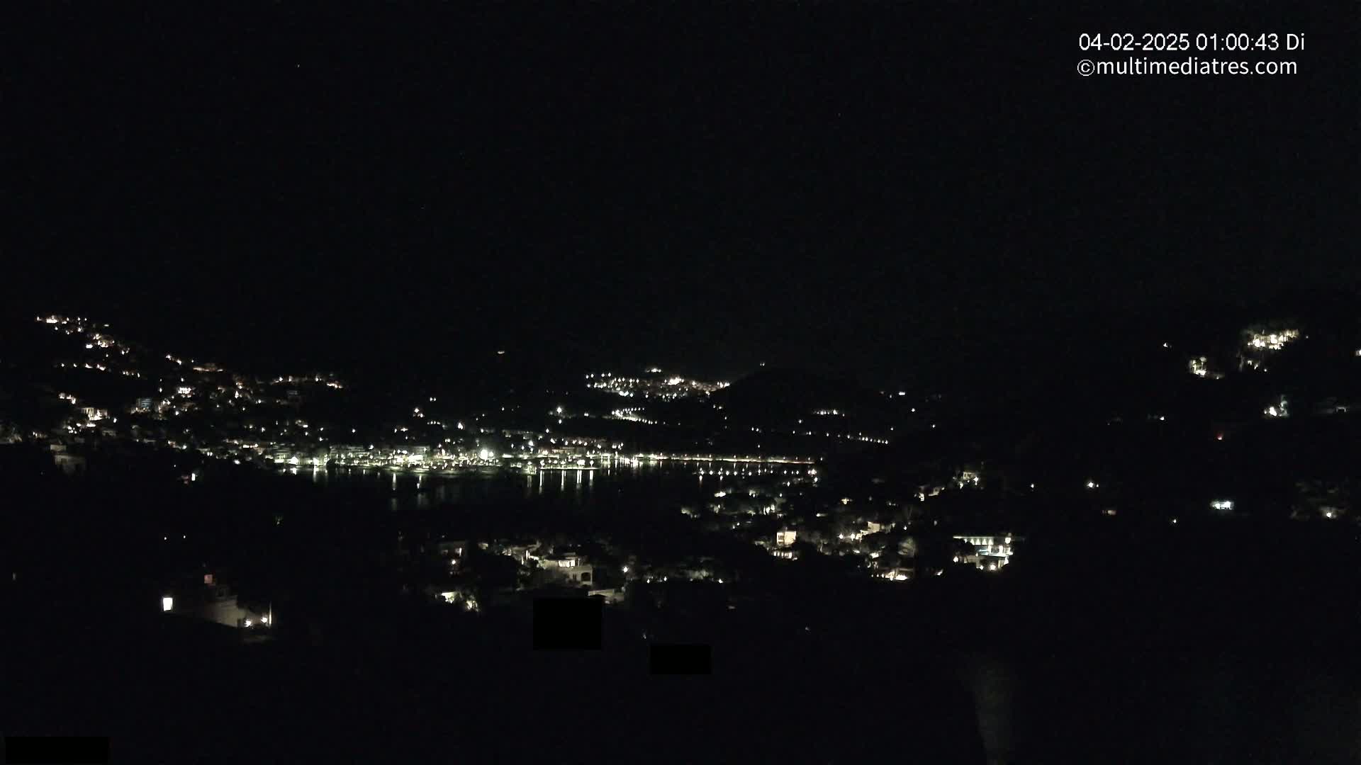 A nighttime view of a coastal town nestled on hillsides, with its lights reflecting on a dark body of water under a clear, starless sky.