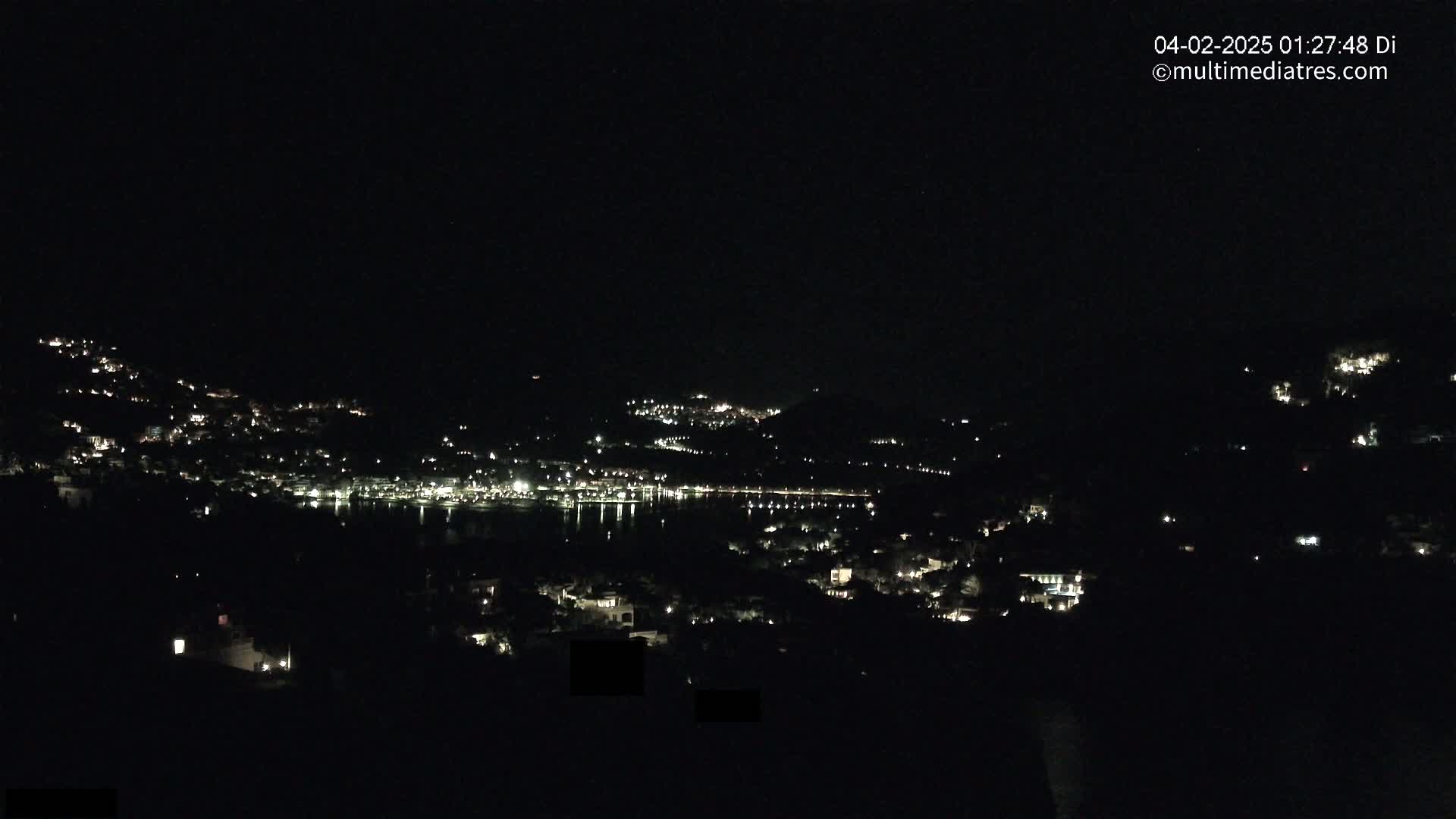 A nighttime view of a coastal town with scattered lights on hillsides and along a dark body of water.