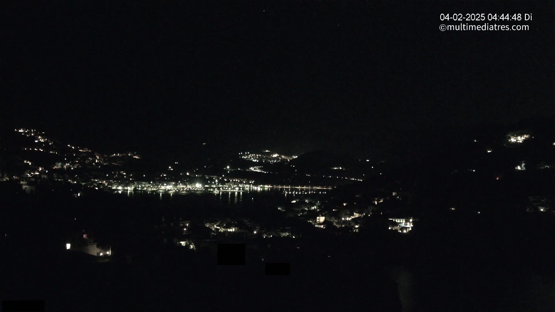 A nighttime view shows a town with scattered lights on hillsides surrounding a dark body of water.