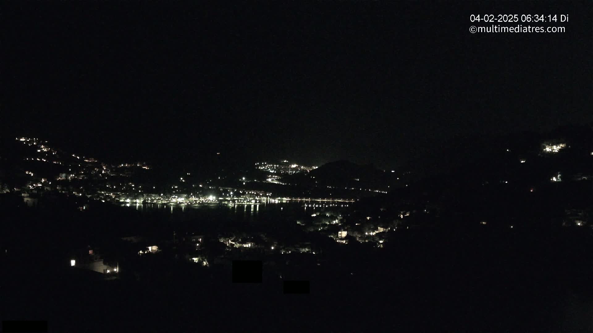 A nighttime view of a town situated on a hillside overlooking a body of water, with scattered lights illuminating the landscape under a dark sky.