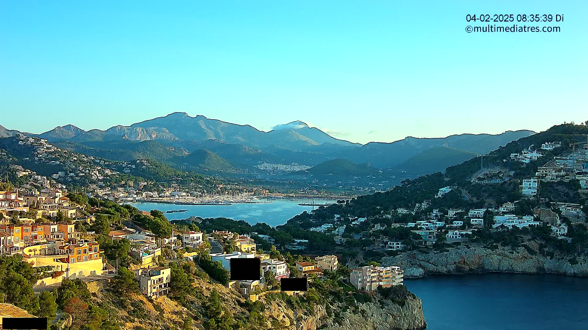 A clear, sunny day reveals a coastal town nestled among hills and mountains overlooking a calm bay filled with boats.