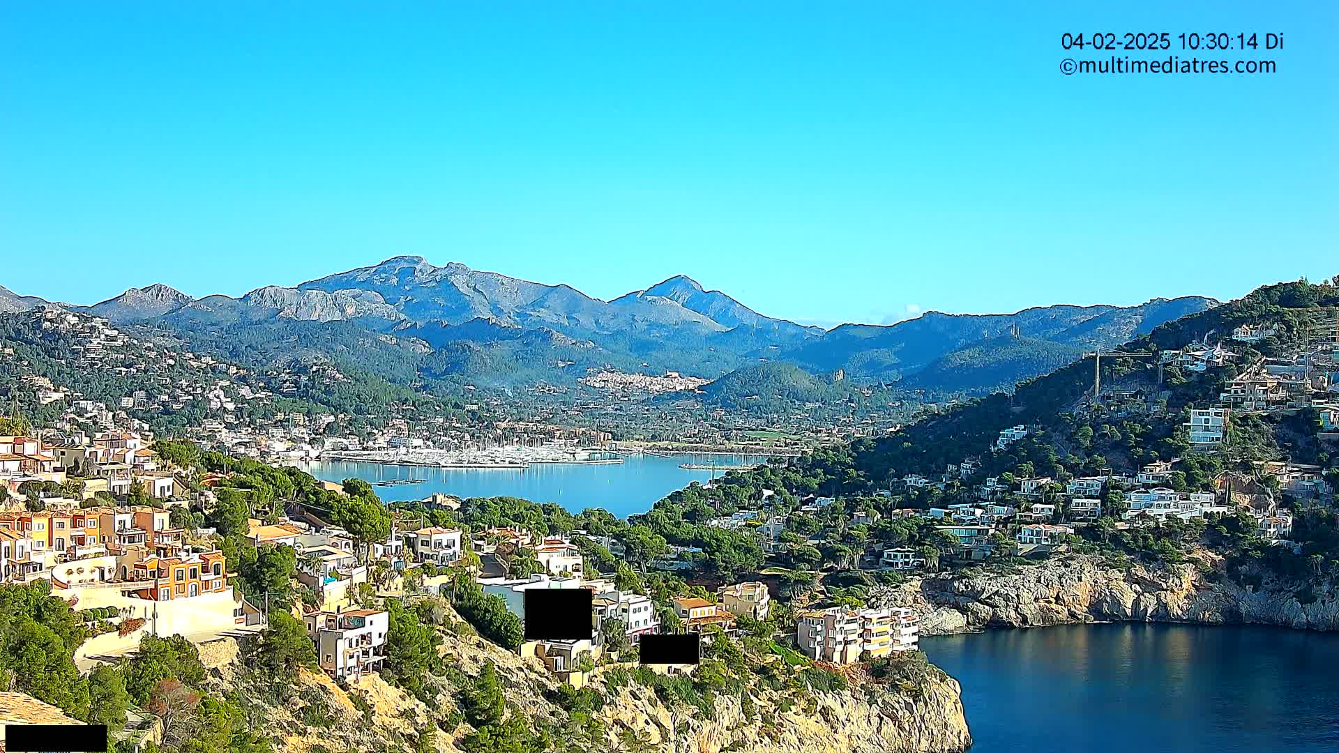A coastal town nestled against a mountain range under a clear blue sky overlooks a calm bay filled with boats.