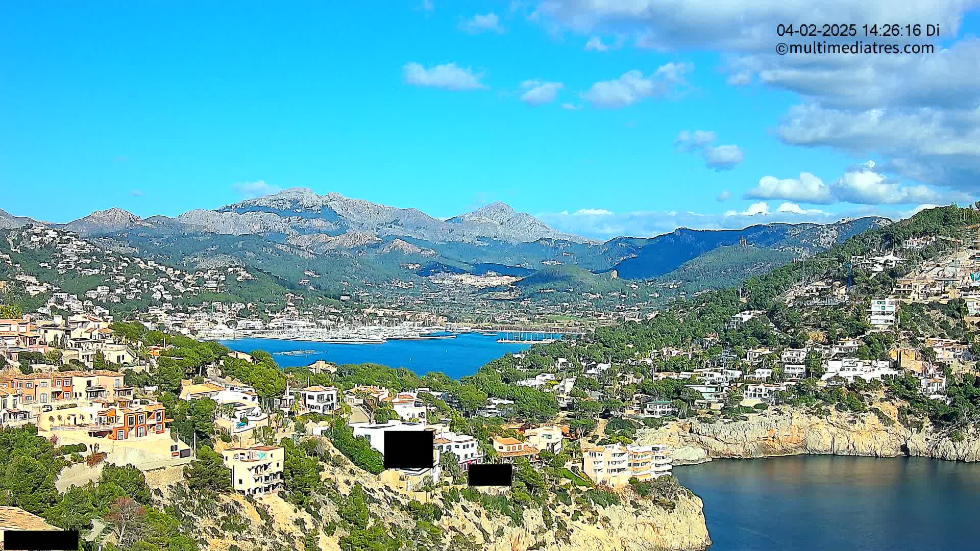 A coastal town nestled into a hillside overlooks a calm bay and mountains under a partly cloudy sky.