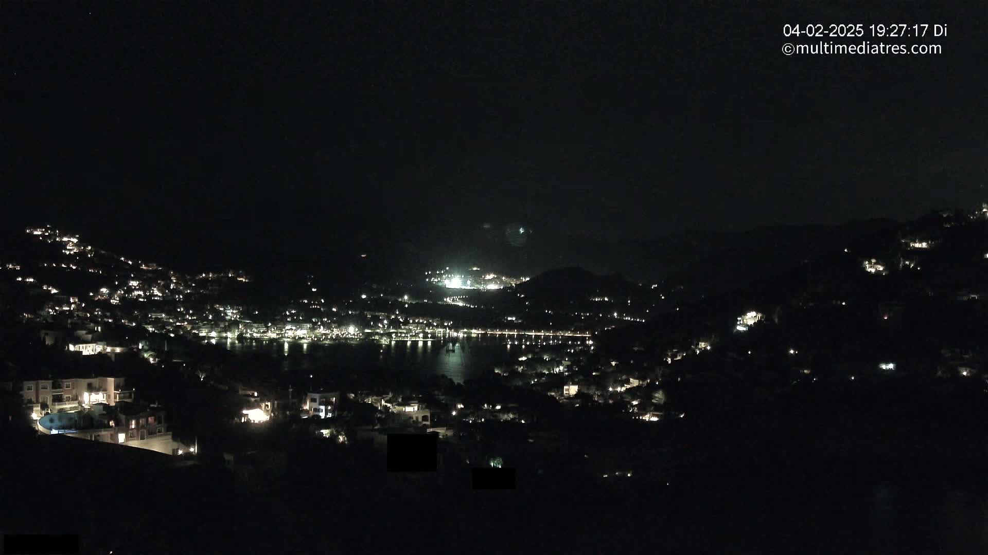 A nighttime view of a coastal town nestled on hillsides, with its lights illuminating the dark landscape and a calm body of water visible in the distance.