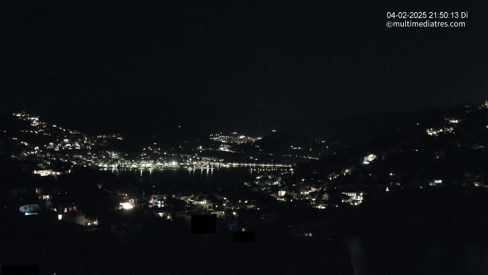 A nighttime view of a coastal town nestled on hillsides, with scattered lights illuminating the buildings and a dark body of water visible in the distance under a clear, dark sky.