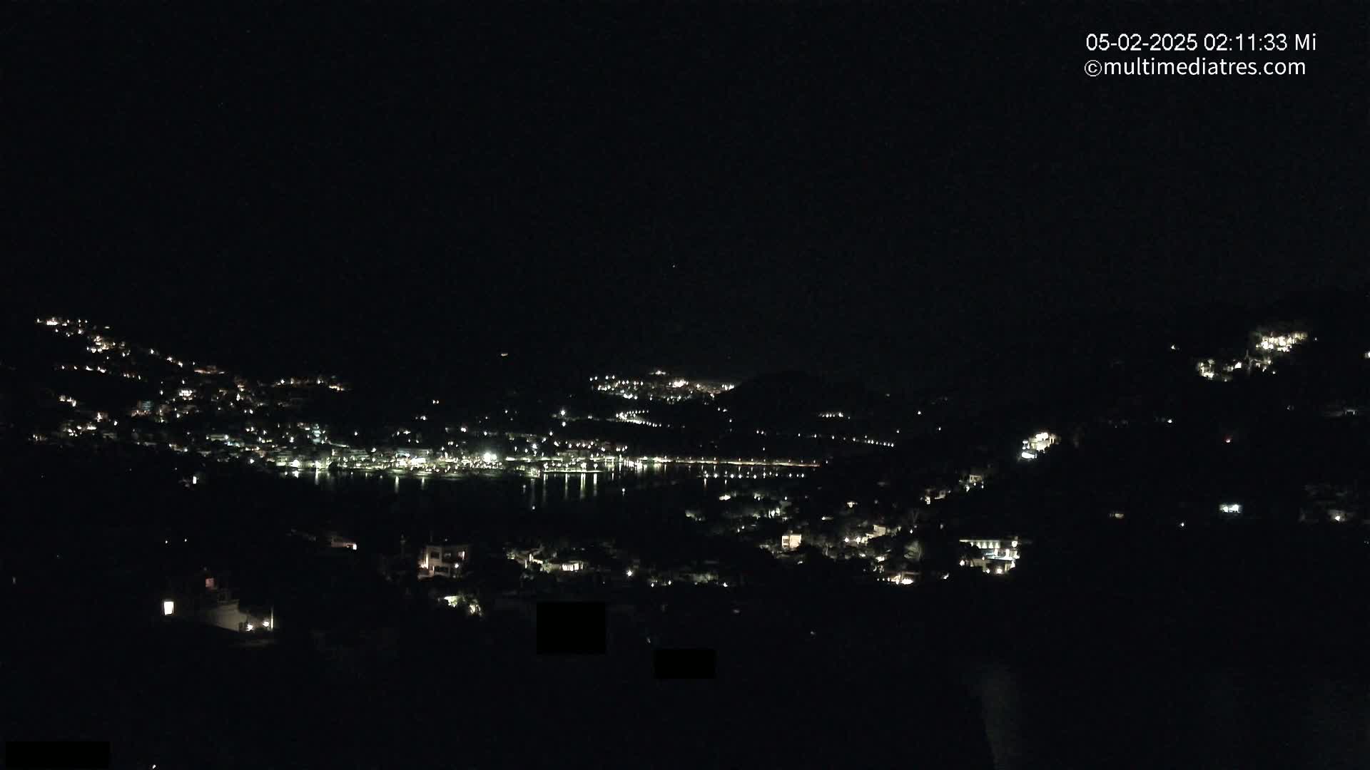 A nighttime view of a town and body of water, with scattered lights illuminating the hillsides under a dark sky.