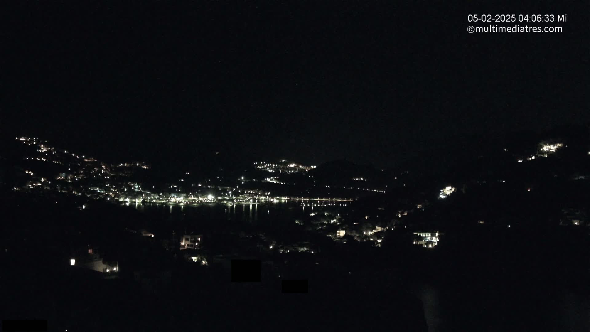 A nighttime view of a town nestled on hillsides surrounding a dark body of water, with scattered lights illuminating the buildings and landscape under a clear, dark sky.