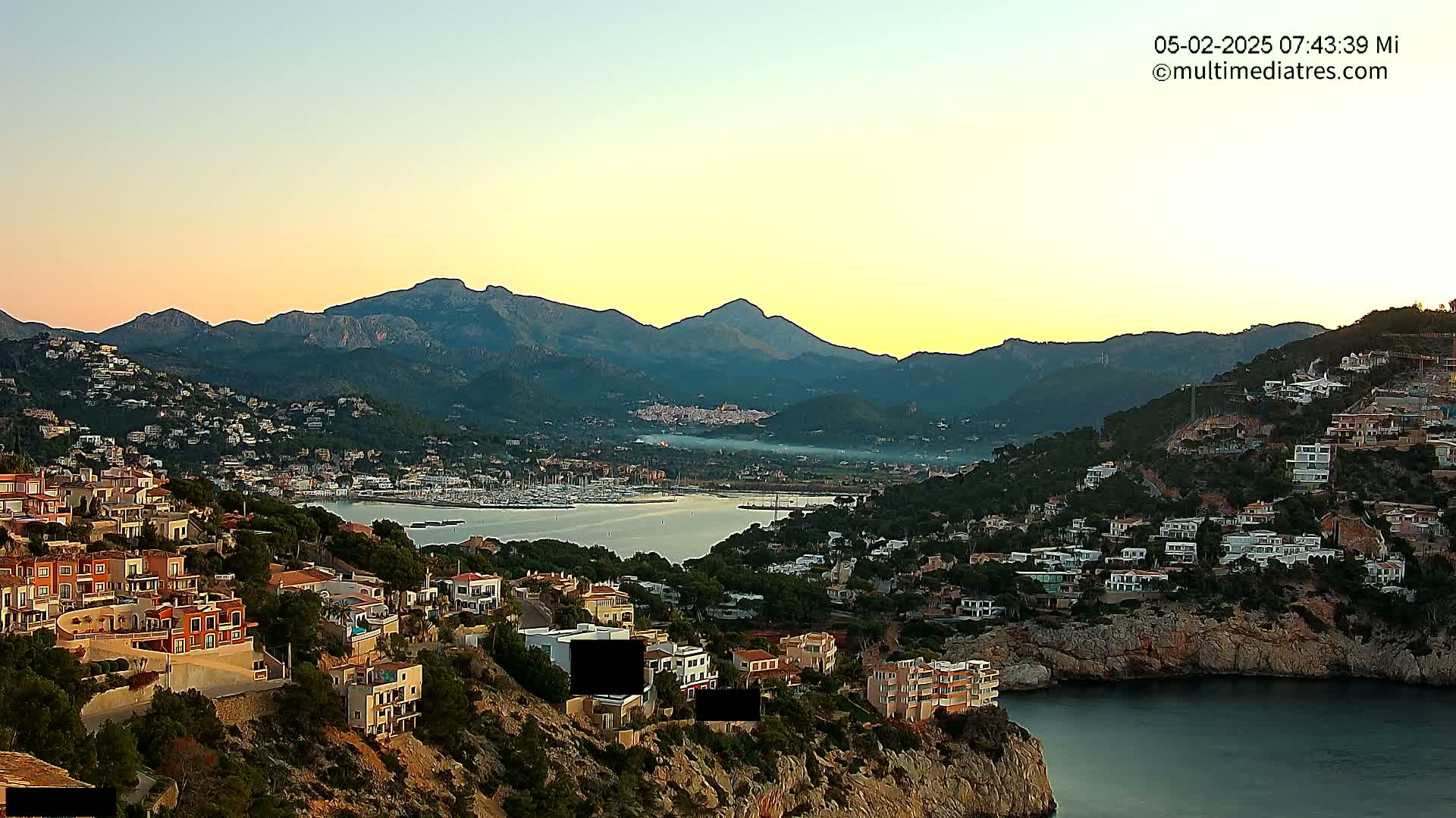 A coastal town nestled in a mountainous region during sunrise, with calm waters in a bay filled with boats.