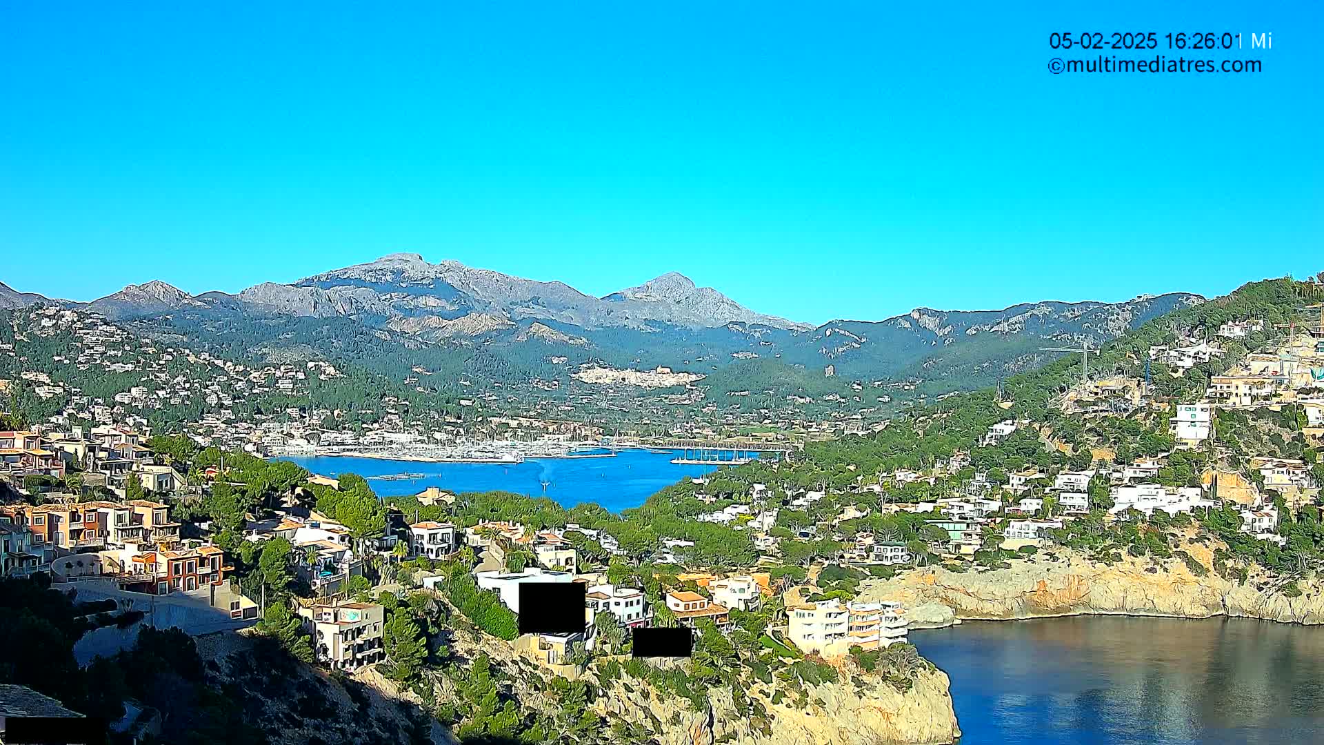 A coastal town nestled against mountains under a clear blue sky overlooks a calm bay filled with boats.