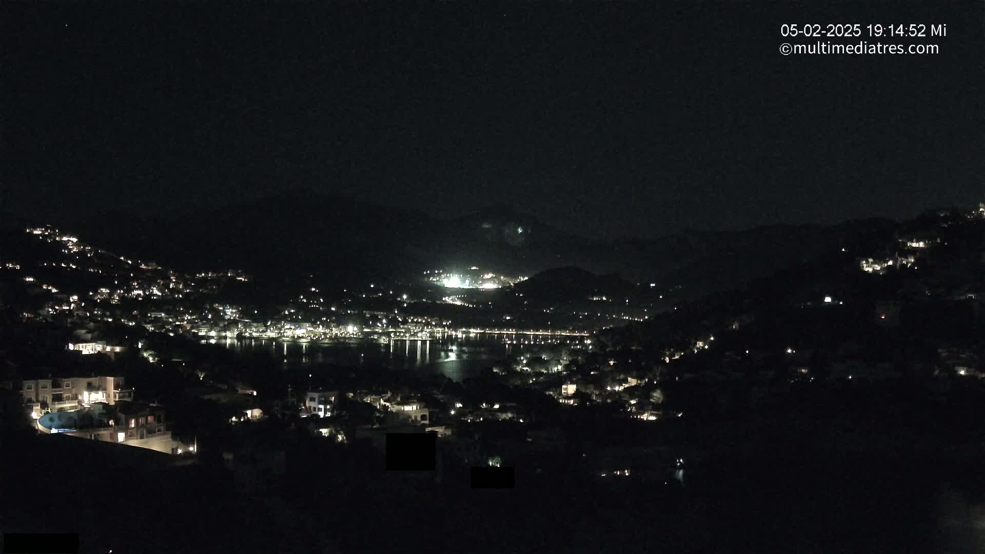 A nighttime view of a coastal town nestled between hills, with numerous lights illuminating buildings and streets along the shoreline and hillsides.