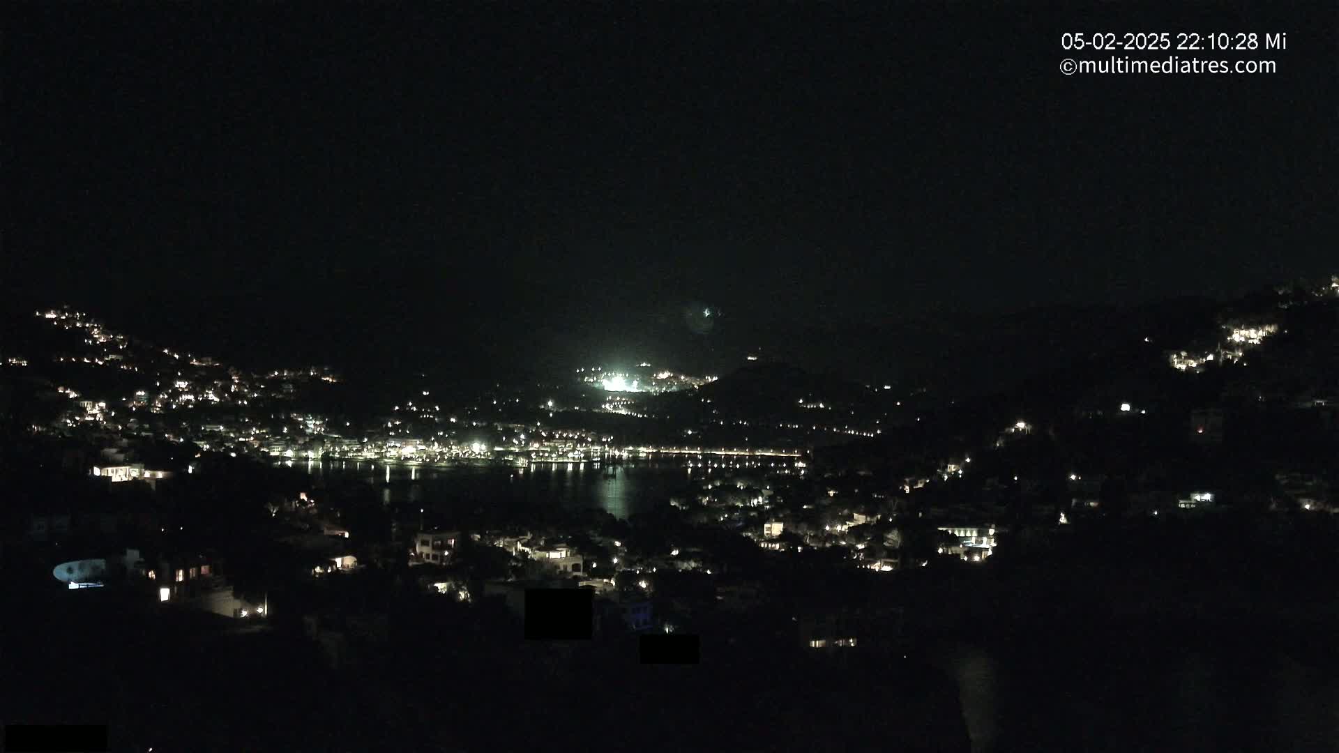 A nighttime view shows a coastal town nestled on hillsides, its buildings illuminated by scattered lights, overlooking a dark body of water under a clear, dark sky.