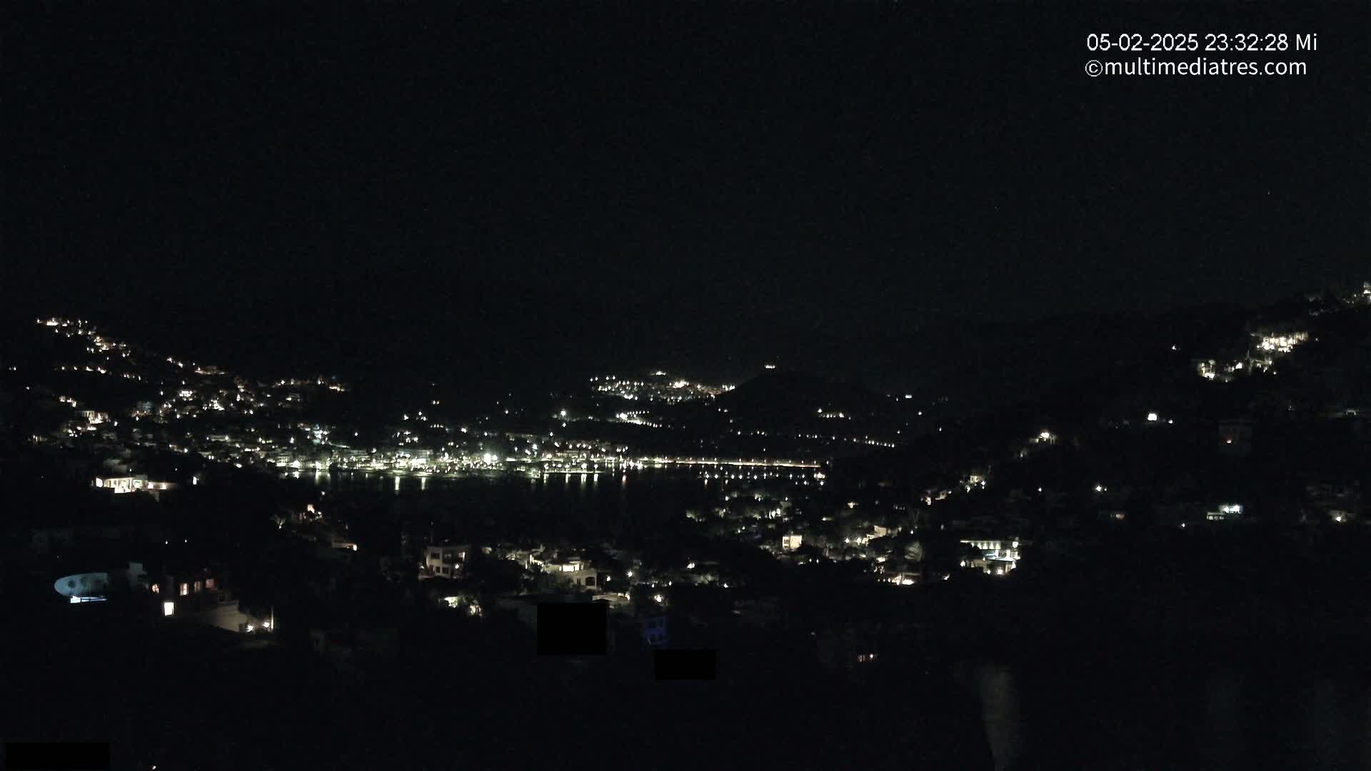 A nighttime view shows a coastal town's lights scattered across hillsides and along a dark body of water under a clear, dark sky.