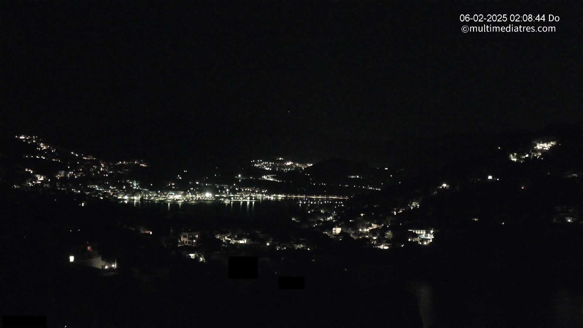 A nighttime view of a town nestled on hillsides around a dark body of water, under a clear sky.
