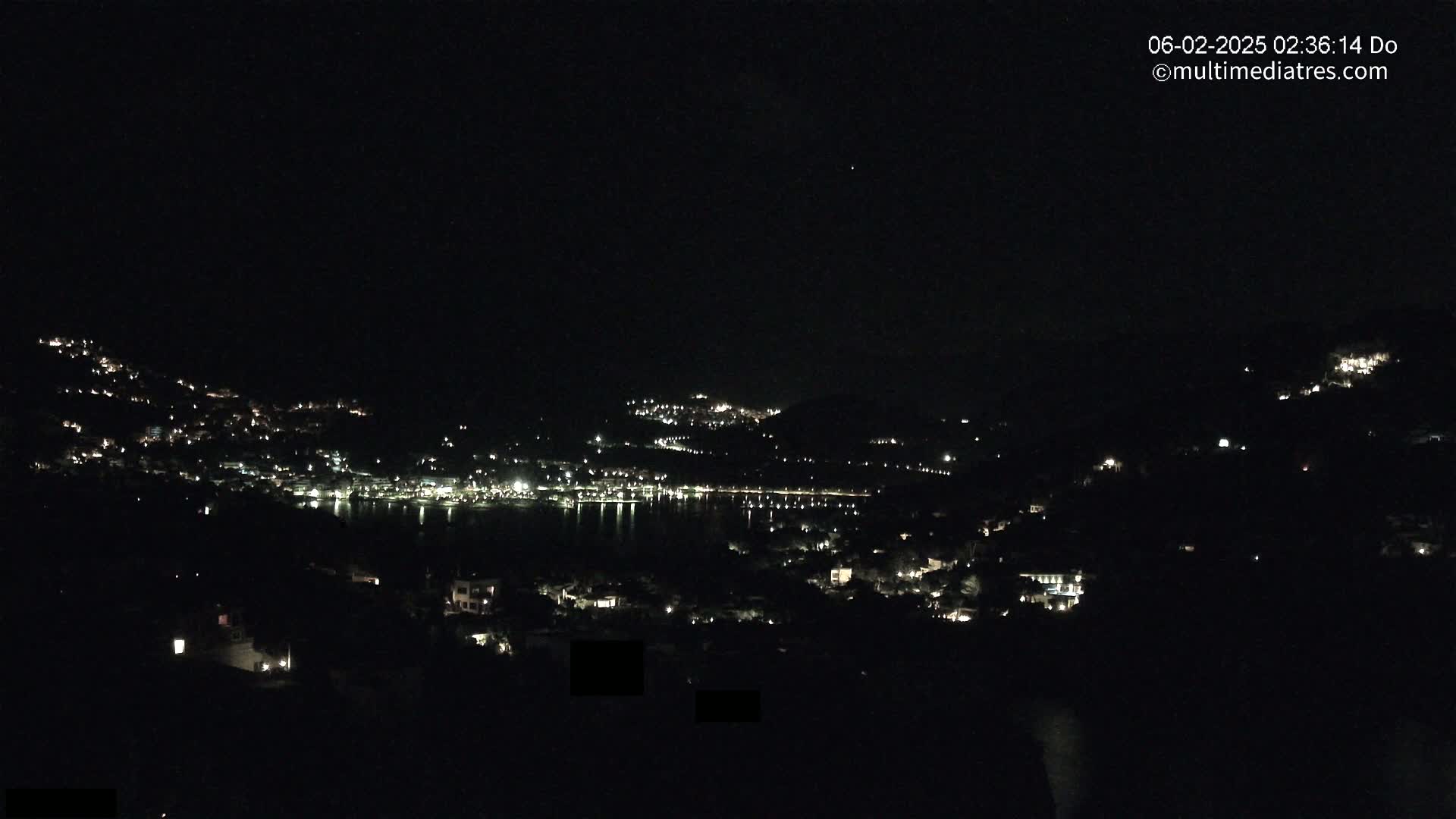 A nighttime view of a coastal town nestled on hillsides, with scattered lights illuminating the buildings and a dark body of water reflecting the lights.