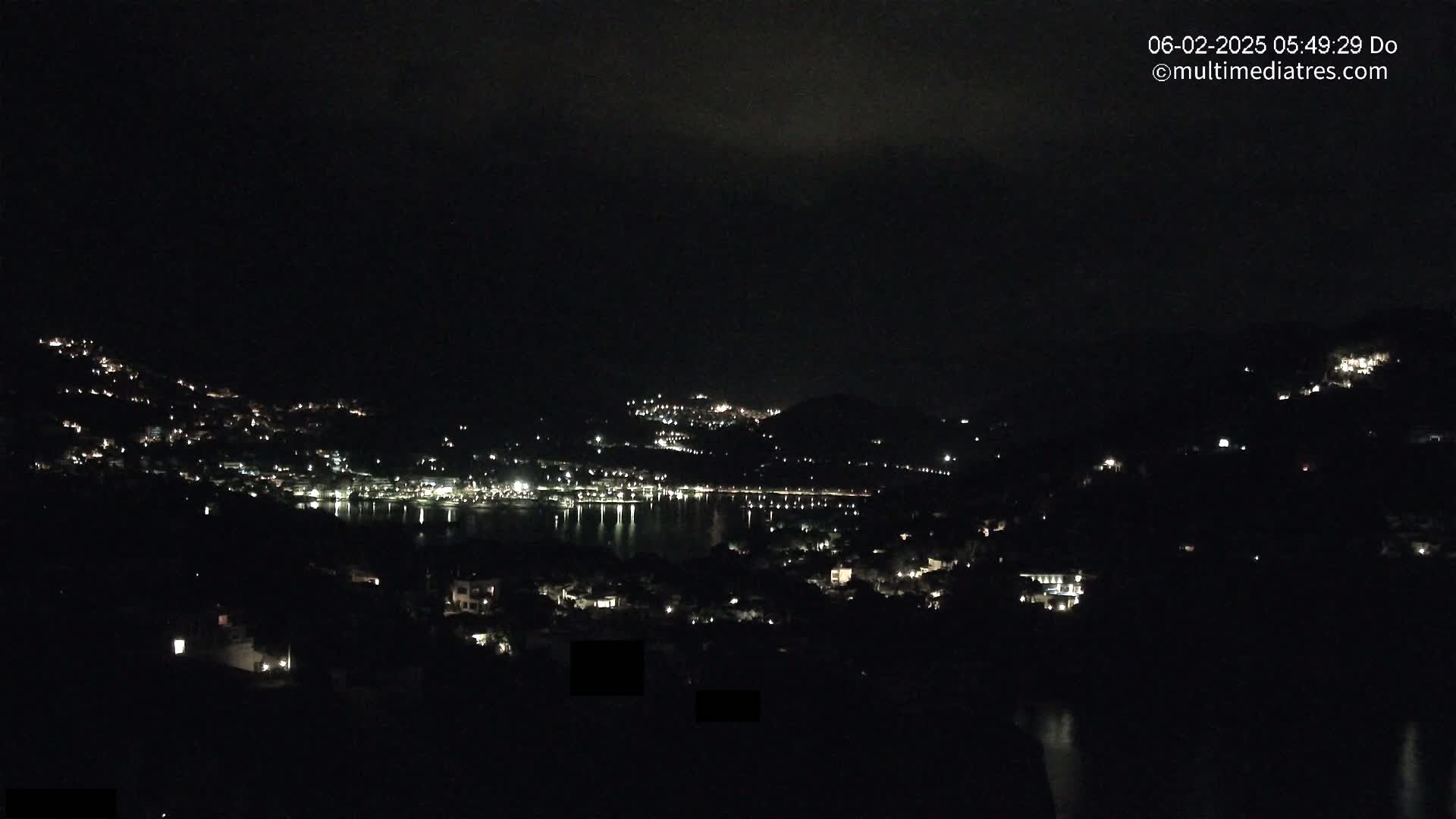 A nighttime view of a coastal town with scattered lights across hillsides and along a calm body of water under a dark, cloudy sky.