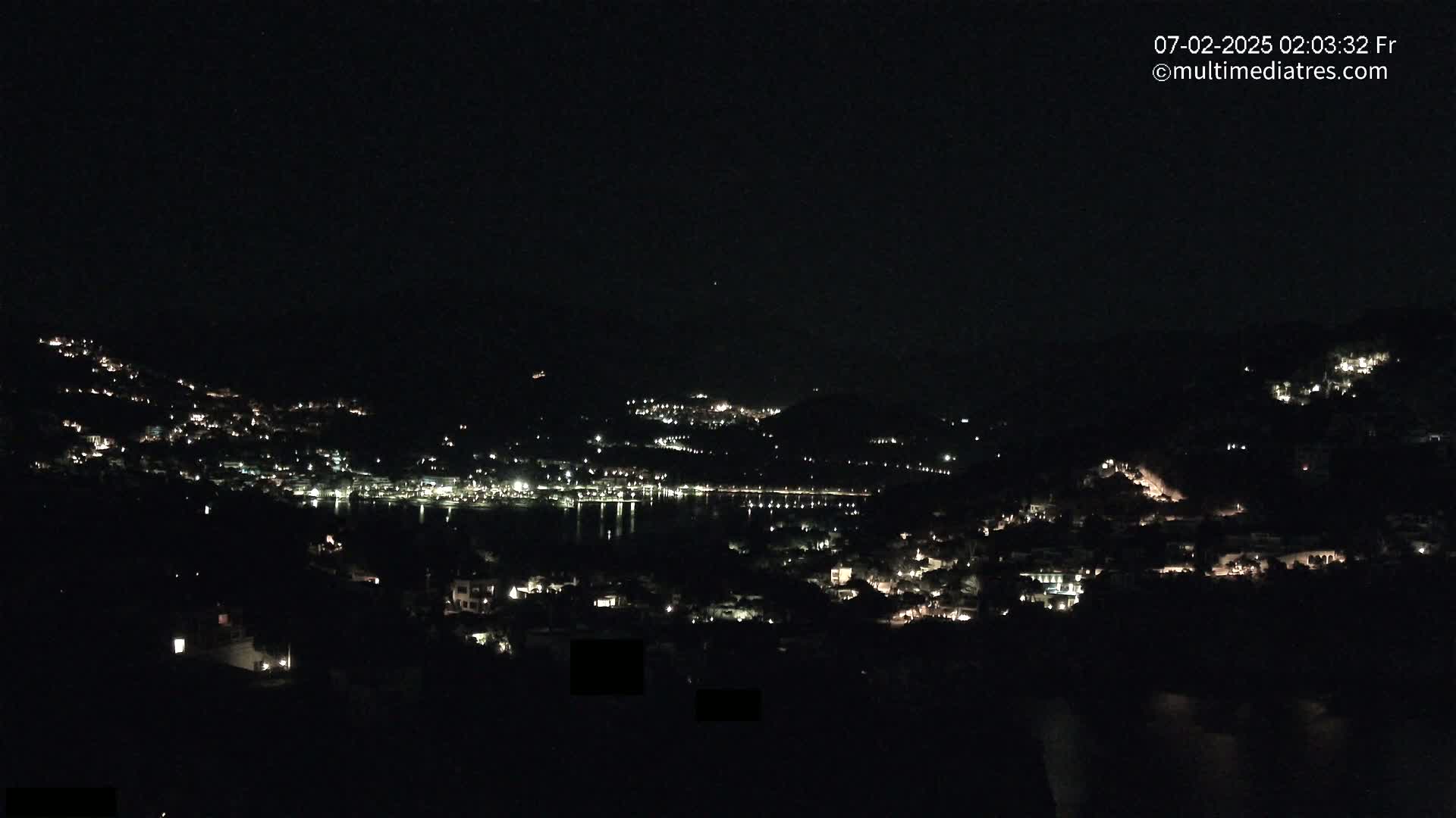 A nighttime view of a town nestled on hillsides surrounding a dark body of water, under a clear, dark sky.