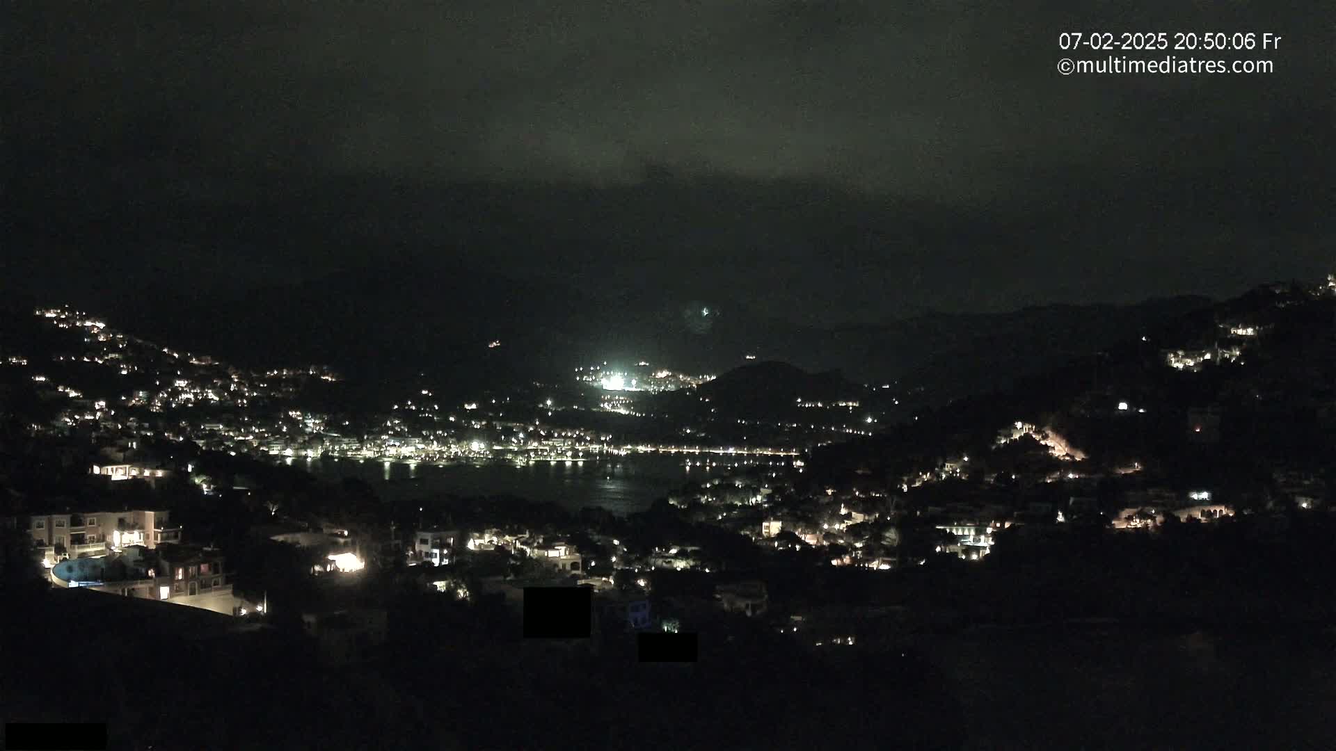 A nighttime view of a coastal town nestled in hills, with many lights illuminating buildings and a dark body of water visible in the distance under a cloudy sky.