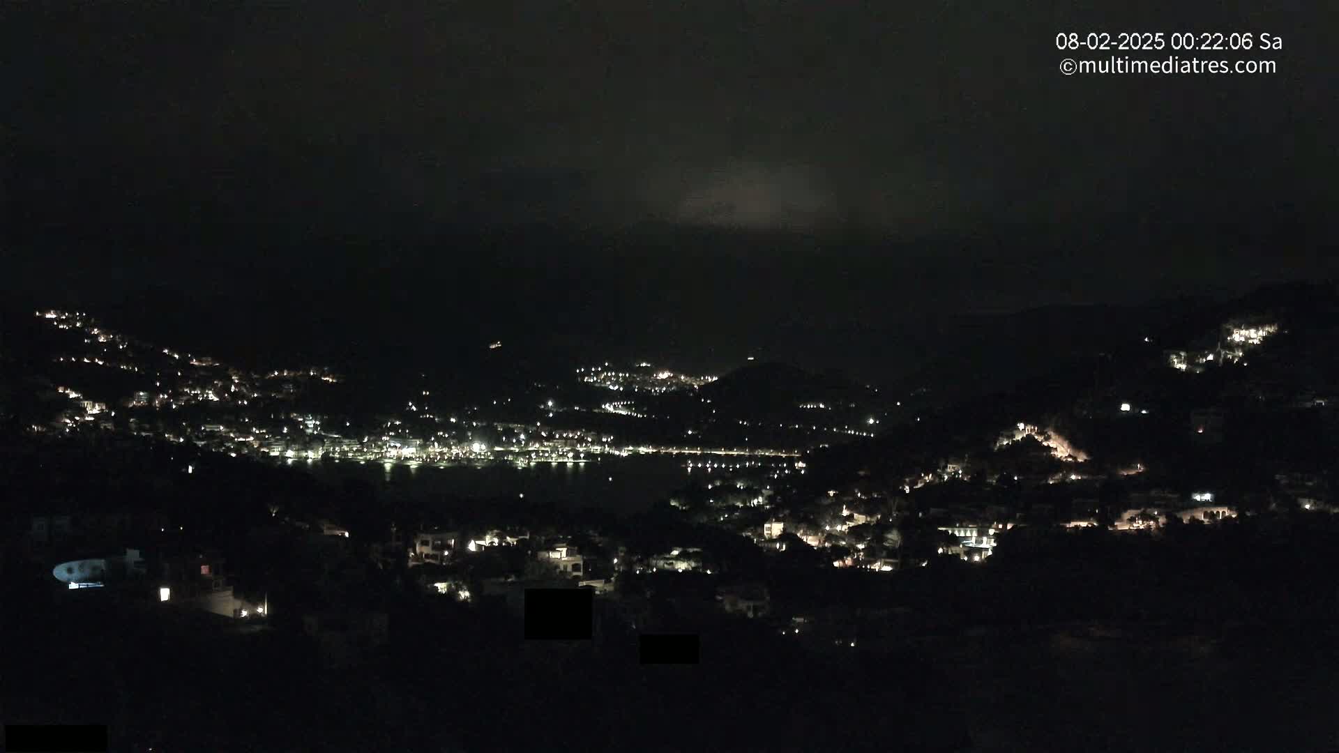 A nighttime view of a town nestled in hills along a dark body of water under a cloudy sky.