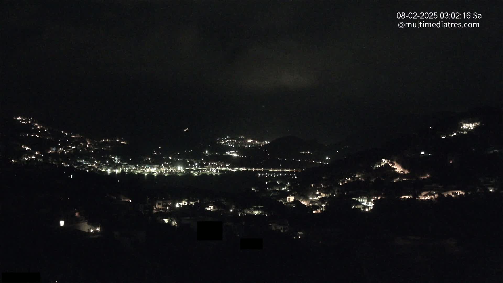 A nighttime view of a hillside town with scattered lights under a dark, cloudy sky.