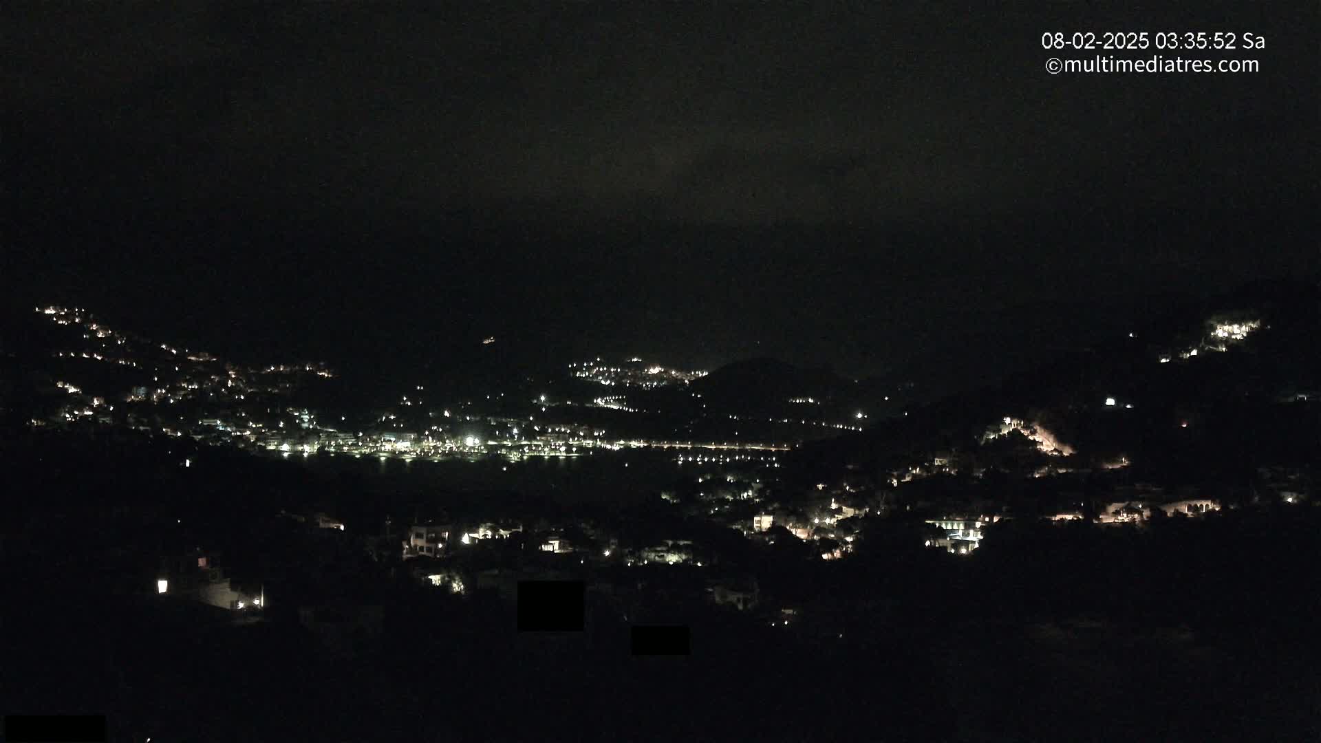 A nighttime, hilltop view of a town with scattered lights under a dark, cloudy sky.